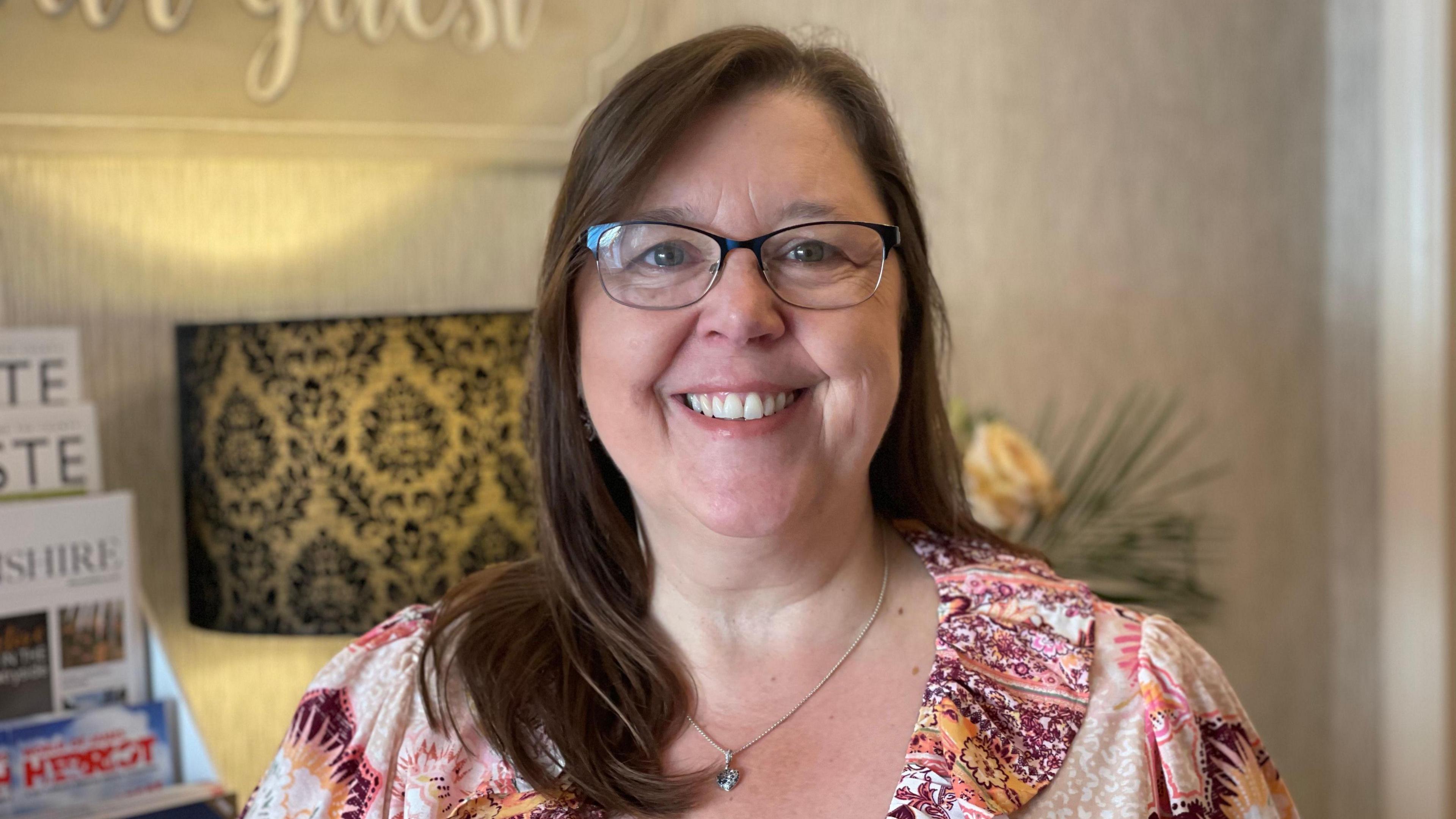 Guest house owner Joanne Veseli has long, dark-brown hair and is smiling while wearing a pink, orange and purple flowery top and dark-rimmed glasses.