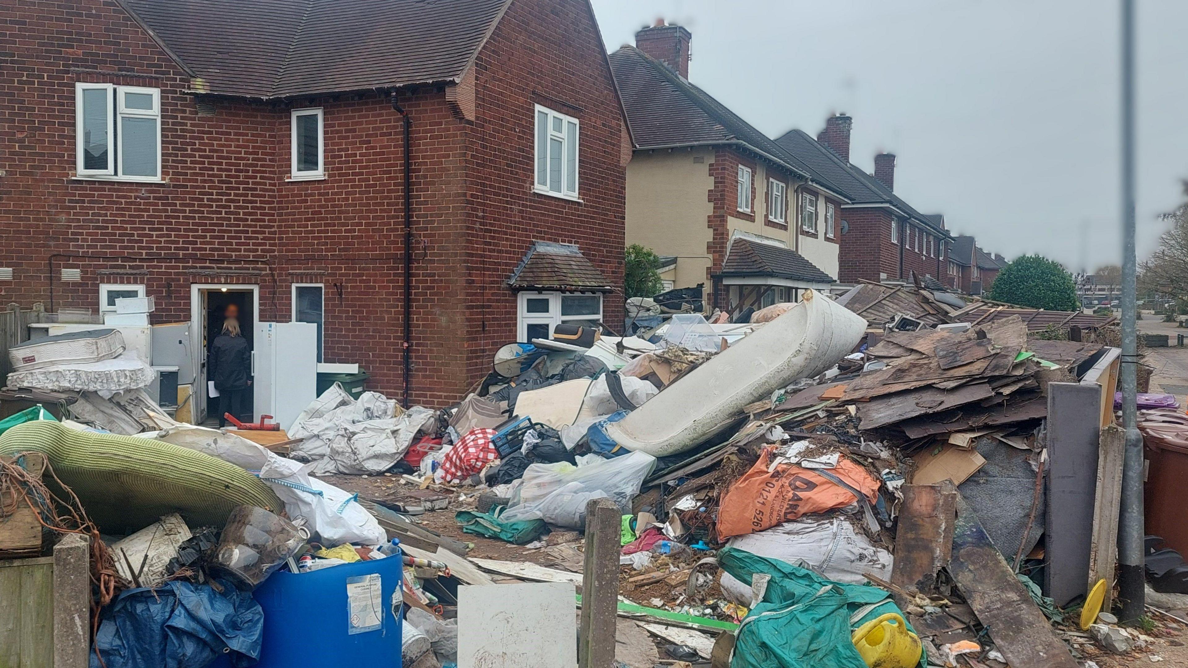 A huge pile of rubbish in front of a house - the rubbish includes bags, furniture, wooden panels and other debris. A woman can be seen in a doorway in the background speaking to someone inside.