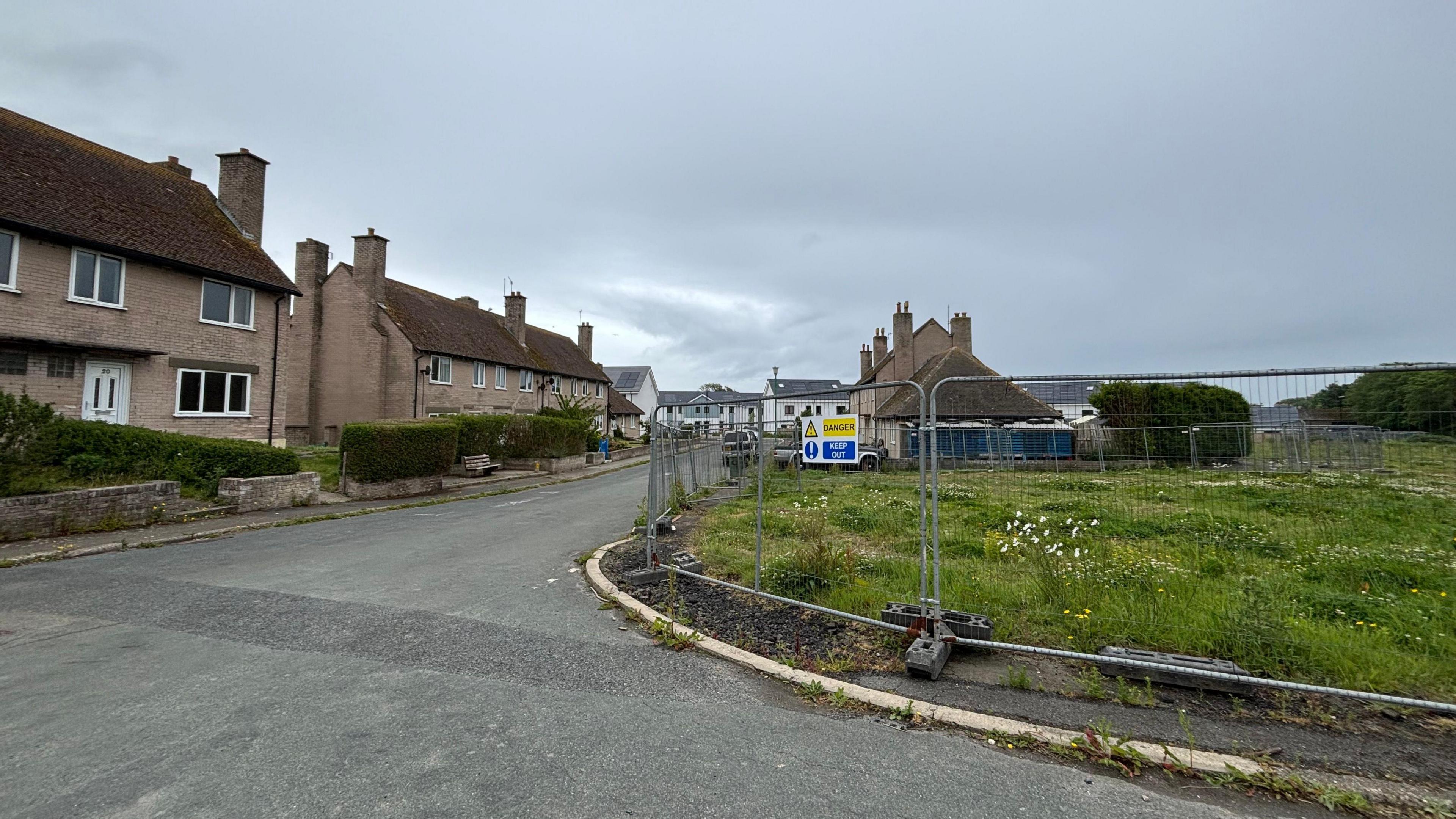 The housing estate as it is now, it is run down, old brown houses are on a road, there is a grassy green space on the right.