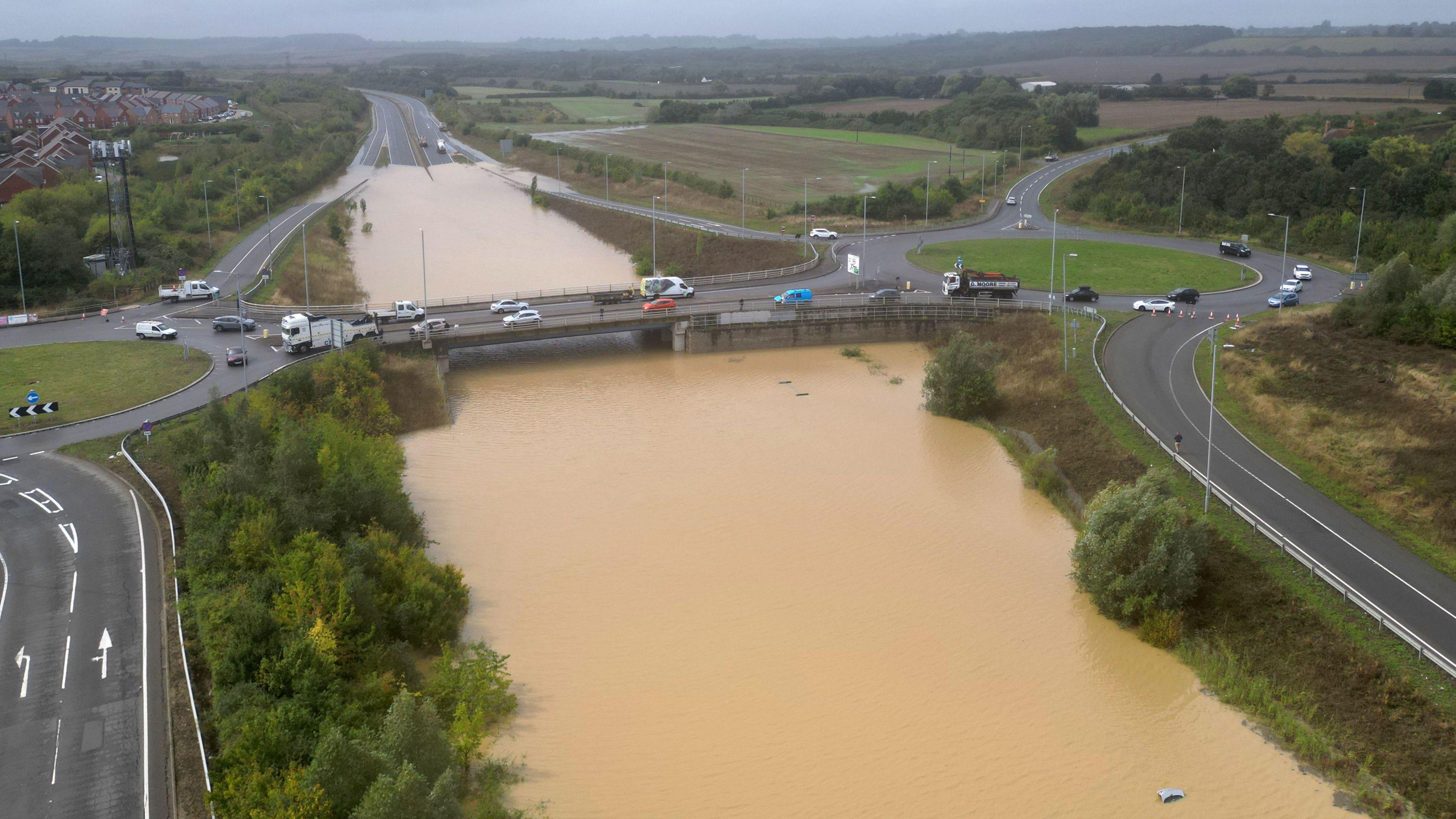 Brown flood water across the main road in Bedfordshire. It nearly reaches the bridge that goes across the stretch of road. You can see a car in the bottom right corner whose boot is sticking up out of the water.