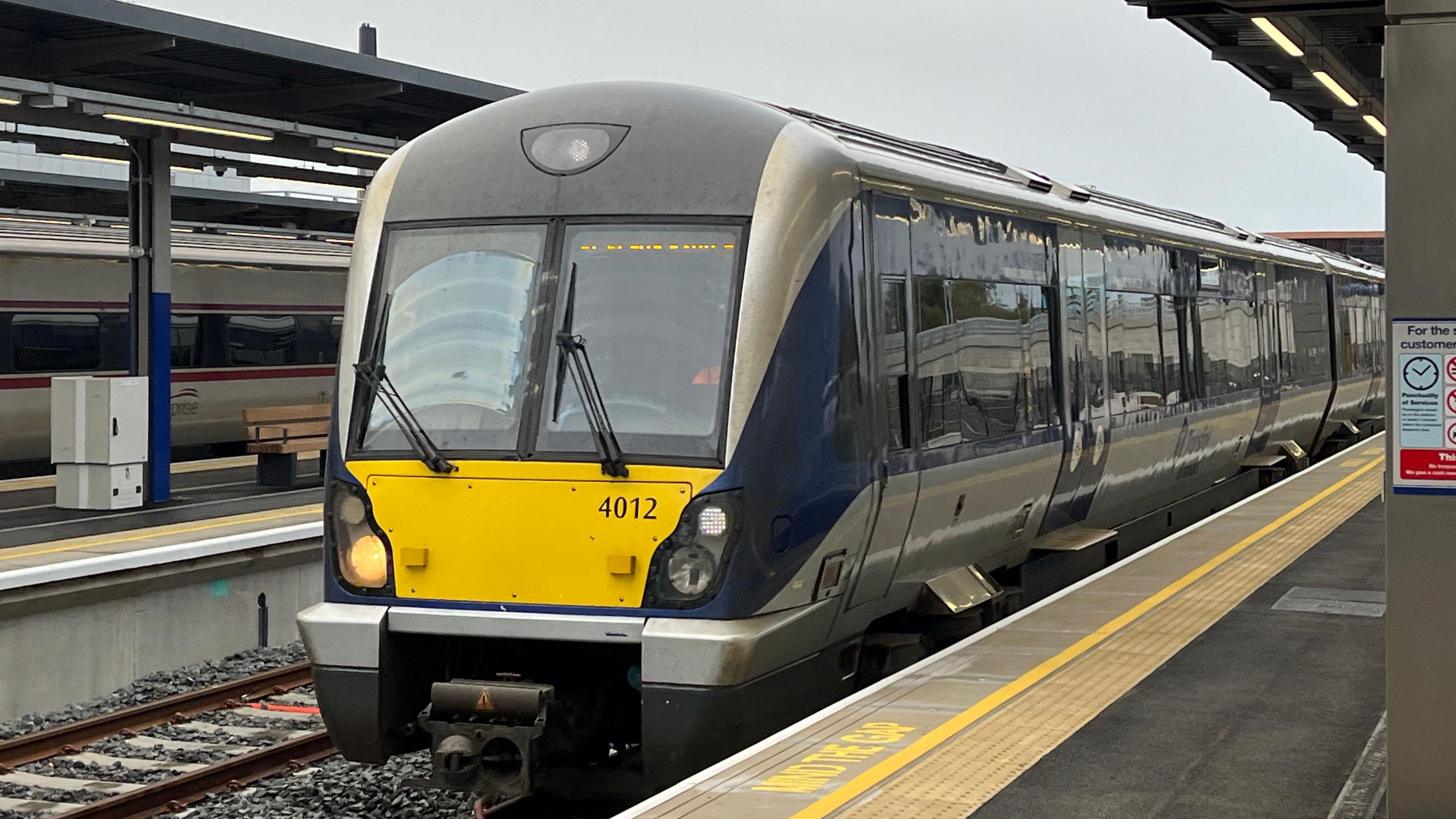 A train is approaching it stop. It is silver yellow and blue. It has the number 4012 in black writing.