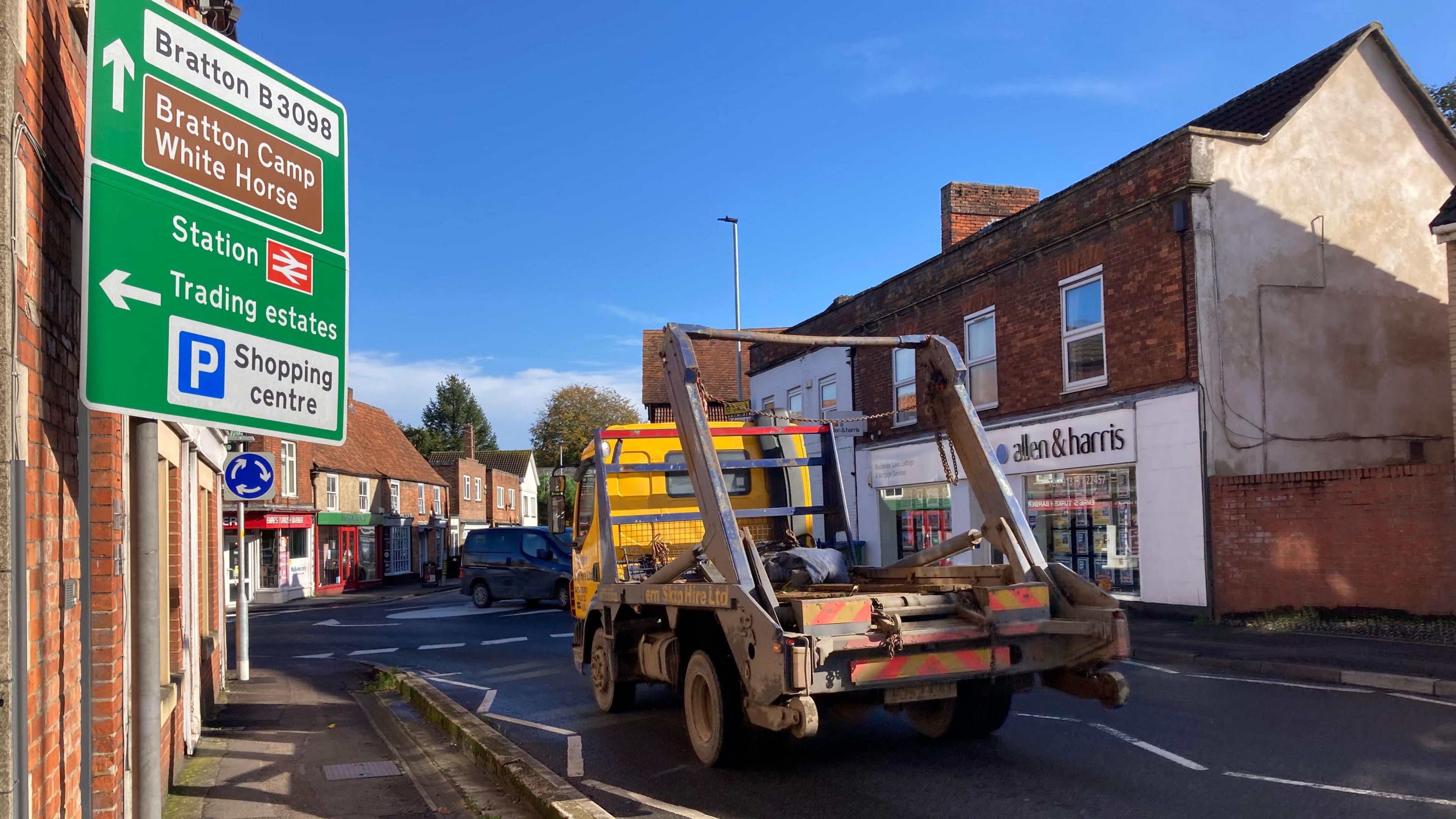 The road going up to a mini roundabout in the middle of Westbury with two-storey red brick buildings and homes on either side, with a skip lorry coming up to the roundabout and a road sign on the left