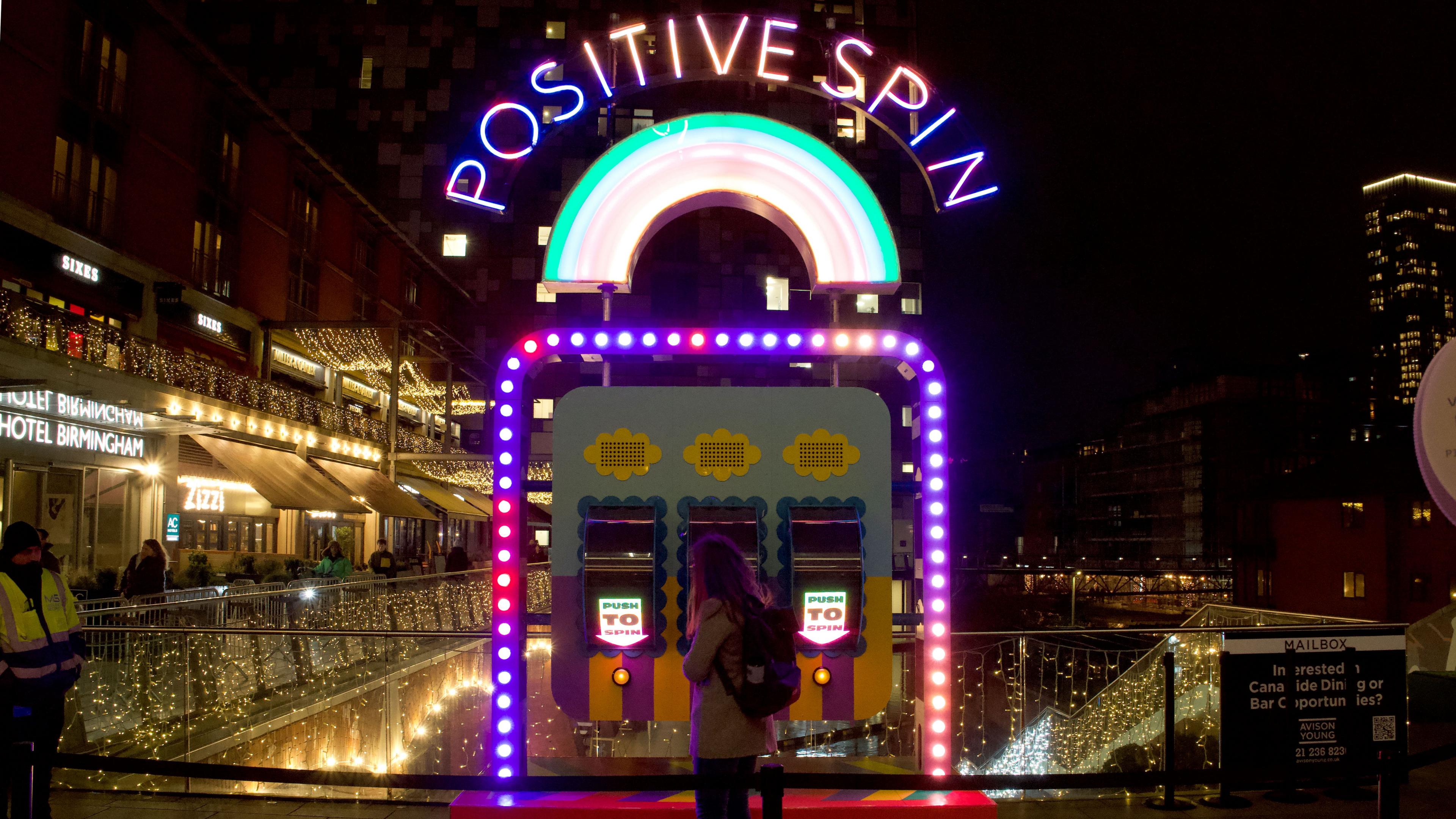 A woman next to a large fruit machine-style installation with three "push to spin" buttons. Above the machine are the words "positive spin"