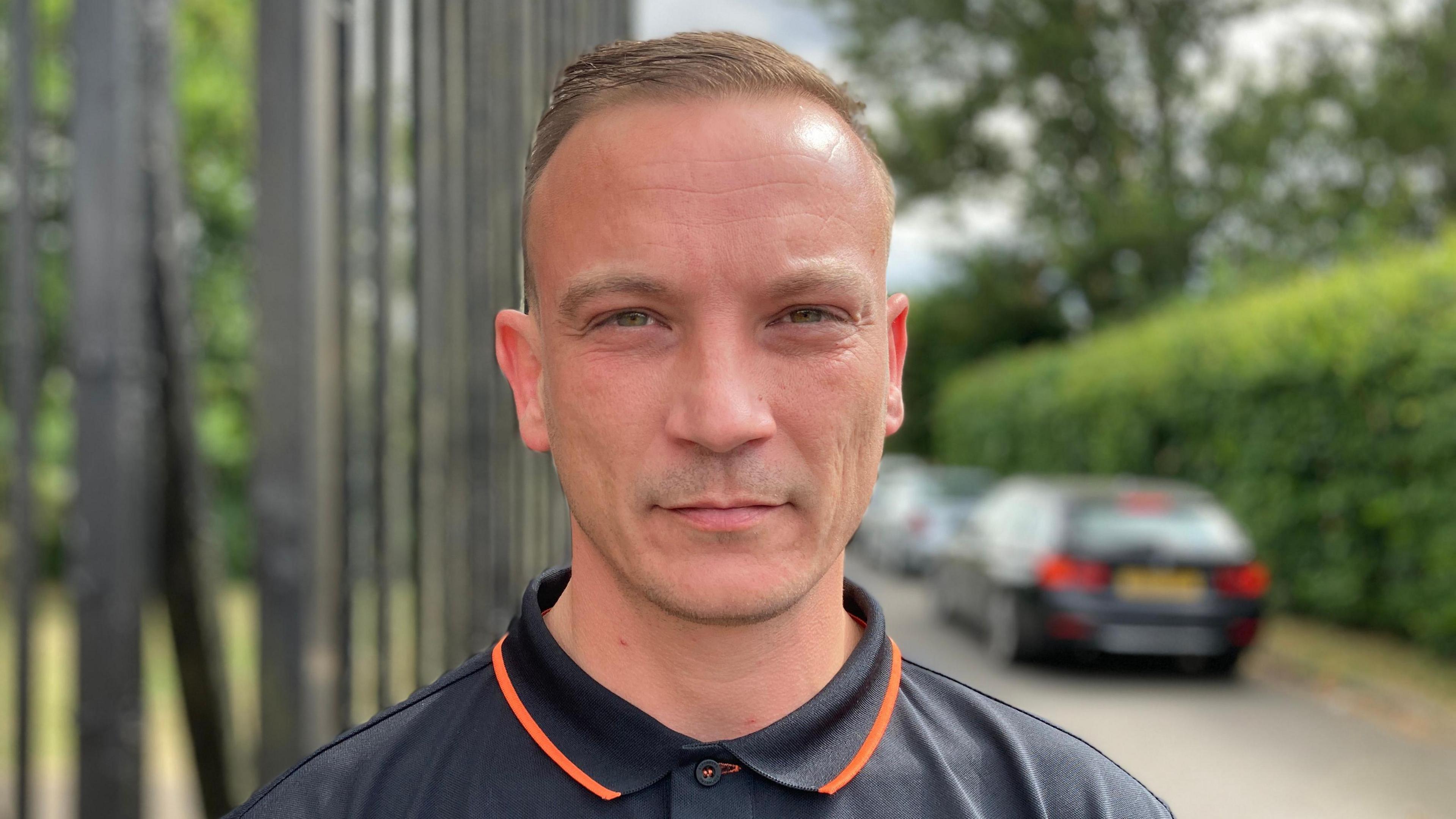 Freddie Cooper is looking directly into the camera, he is wearing a black and orange top. He's standing outside the gates of Saffron Hill cemetery in Leicester.