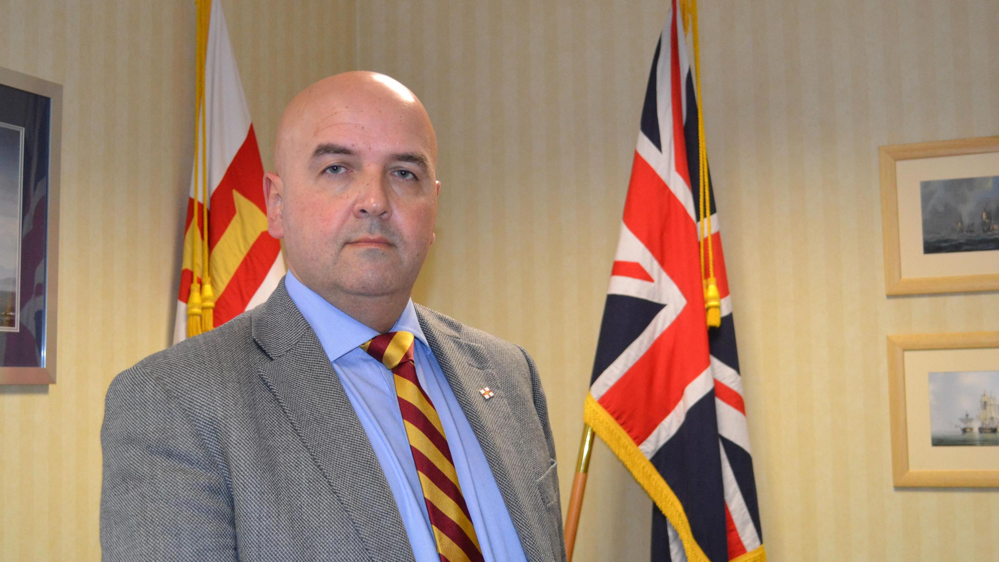 Deputy Jonathan Le Tocq - A man with a bald head, wearing a grey suit, blue button up shirt and a burgundy and yellow tie. He's standing in front of a Union Jack flag and a Guernsey flag.