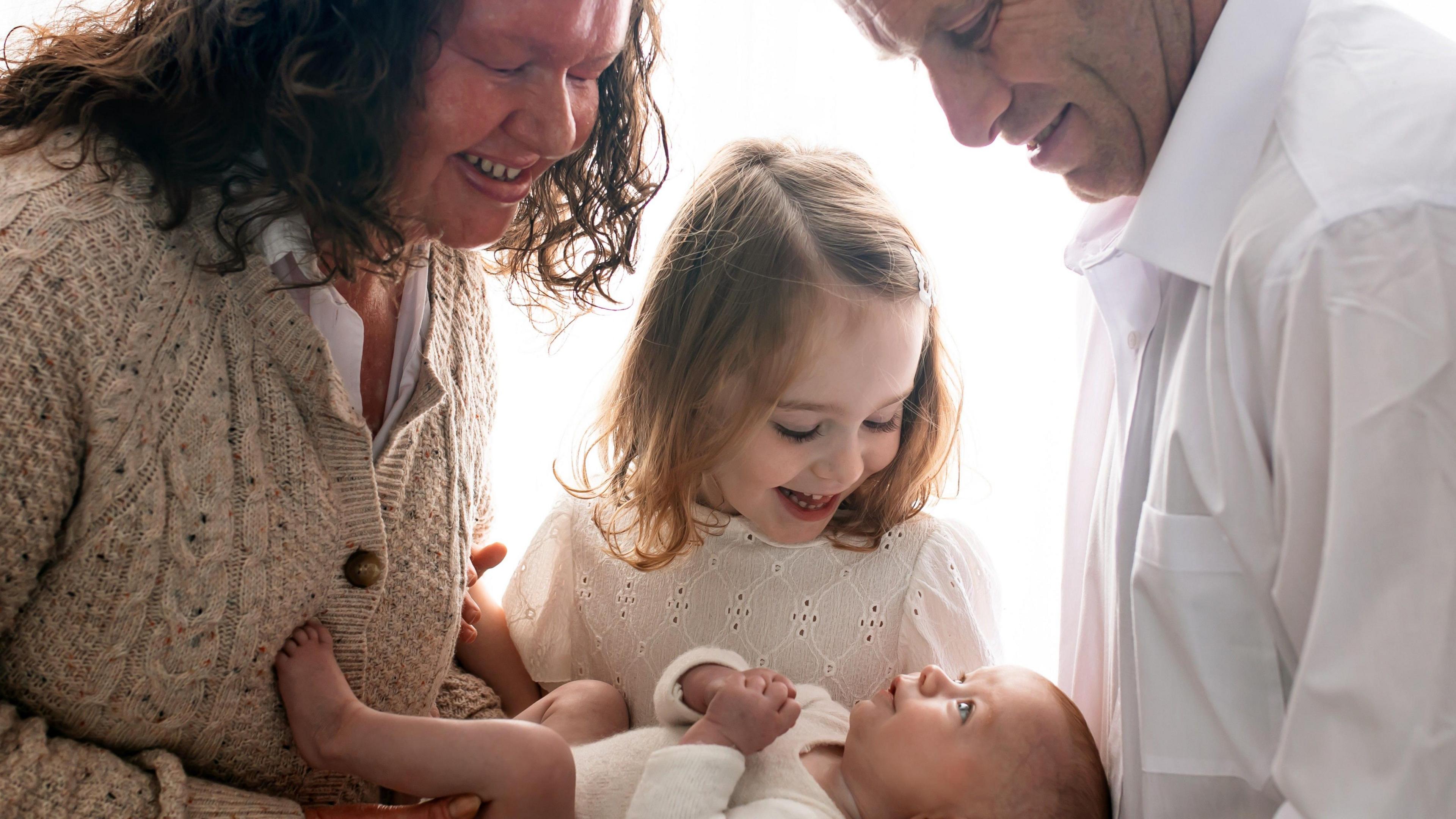 A family portrait showing a mum dressed in an oatmeal cardigan and white shirt with a baby in a white baby grow, a girl in a white top and a man in a white shirt