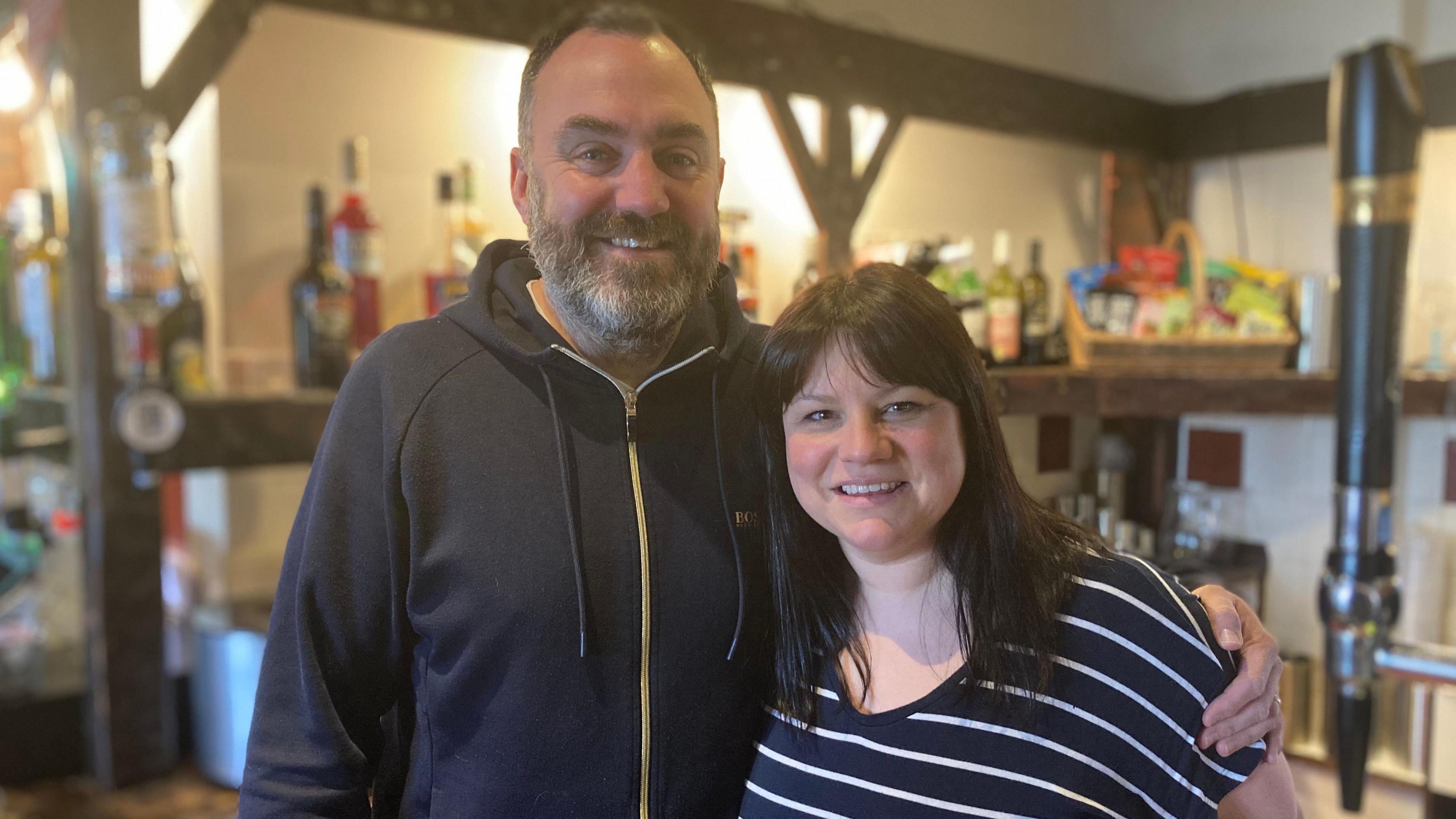 James and Tasmin Kaminiski smile at the camera. James has short brown hair and a greying beard. He is wearing a navy hooded top. Tasmin has ling, dark hair and is wearing a blue and white striped top.