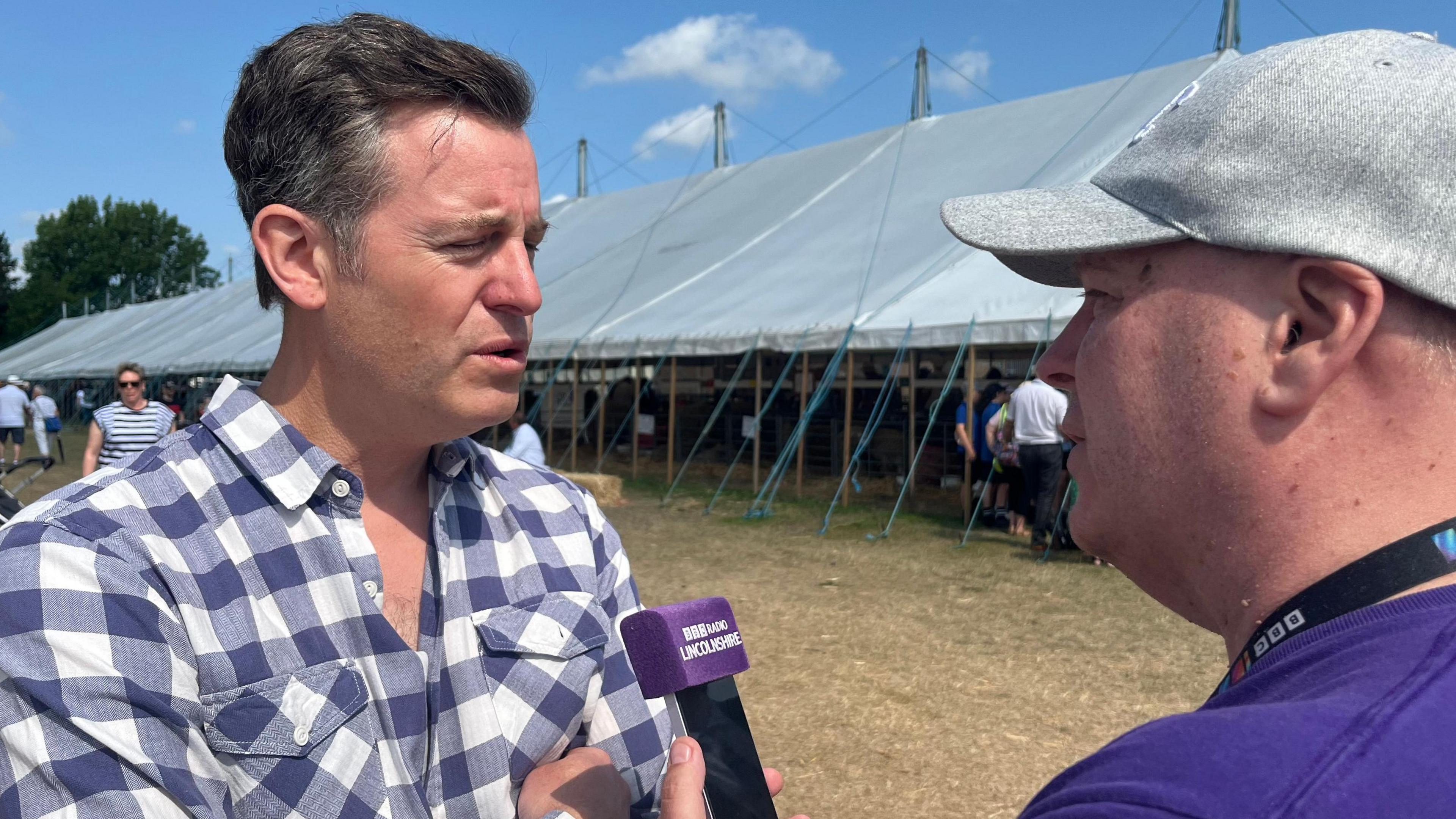 A man wearing a checked shirt being interviewed by another man wearing a grey cap and holding a microphone with the words BBC Radio Lincolnshire on the muff.