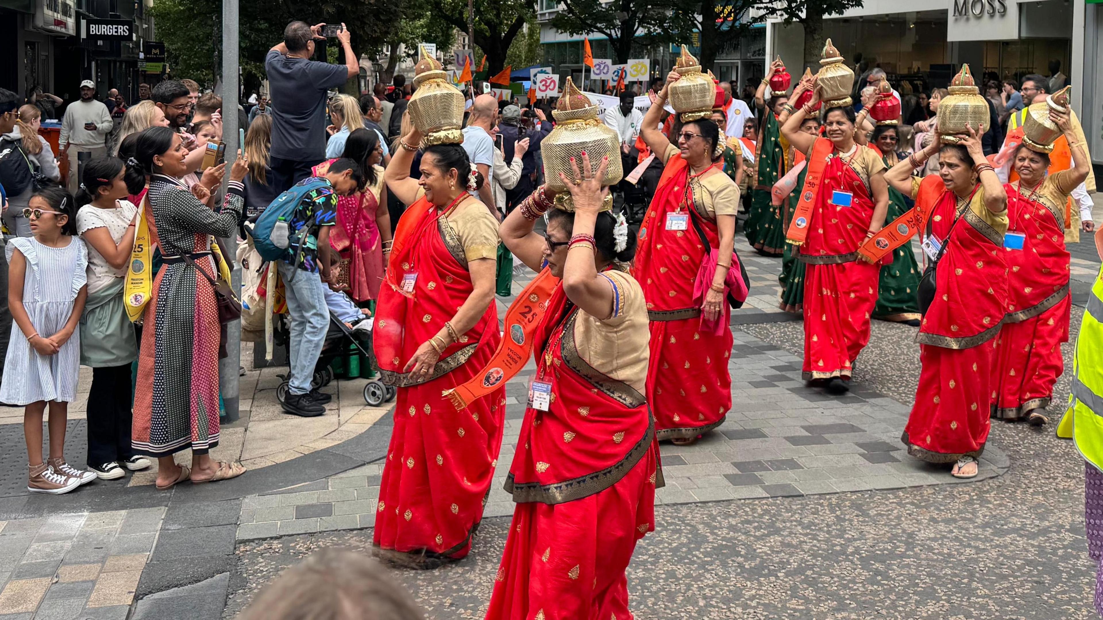 A group of more than six women in red and beige saris hold up golden-coloured vessels on their heads as they walk through a street past spectators, who are watching or taking pictures.