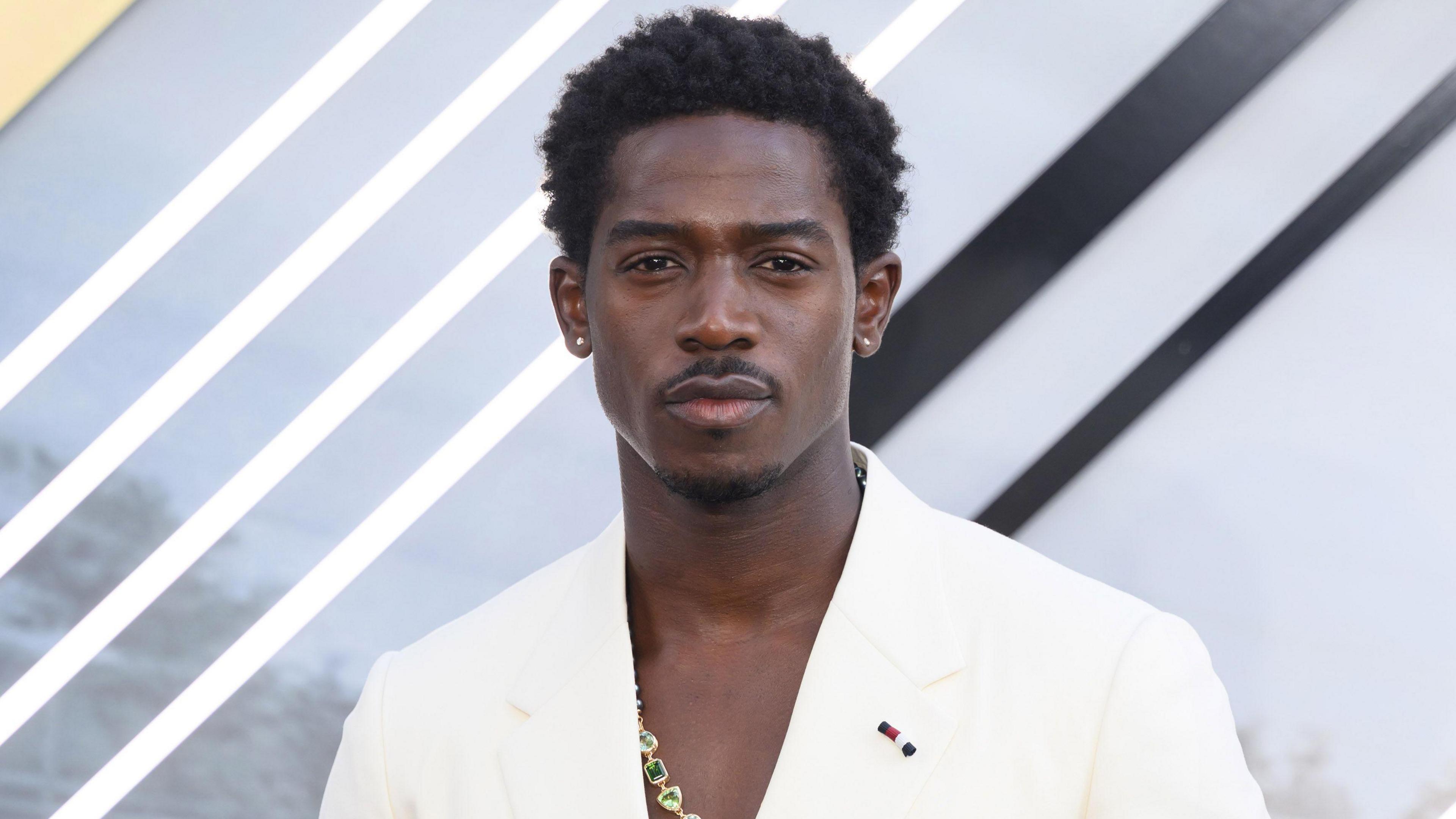 Damson Idris attends the "F1: The Movie" European Premiere at Cineworld Leicester Square on June 23, 2025 in London. The actor has short black hair and a trimmed moustache and goatee. He wears a cream blazer over a bare chest with a gold necklace with green gem stones.