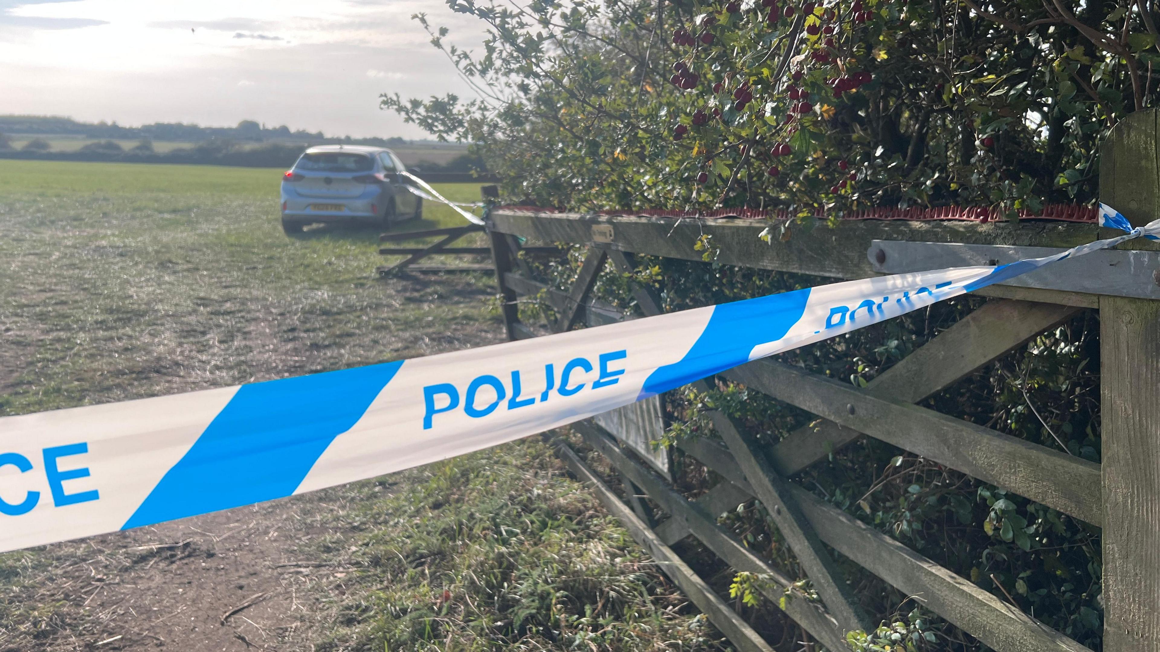 Blue and white tape with the words police written on it stretches across an entrance into a field. It is tied up against a fence pole. A silver car can be seen parked in the field.
