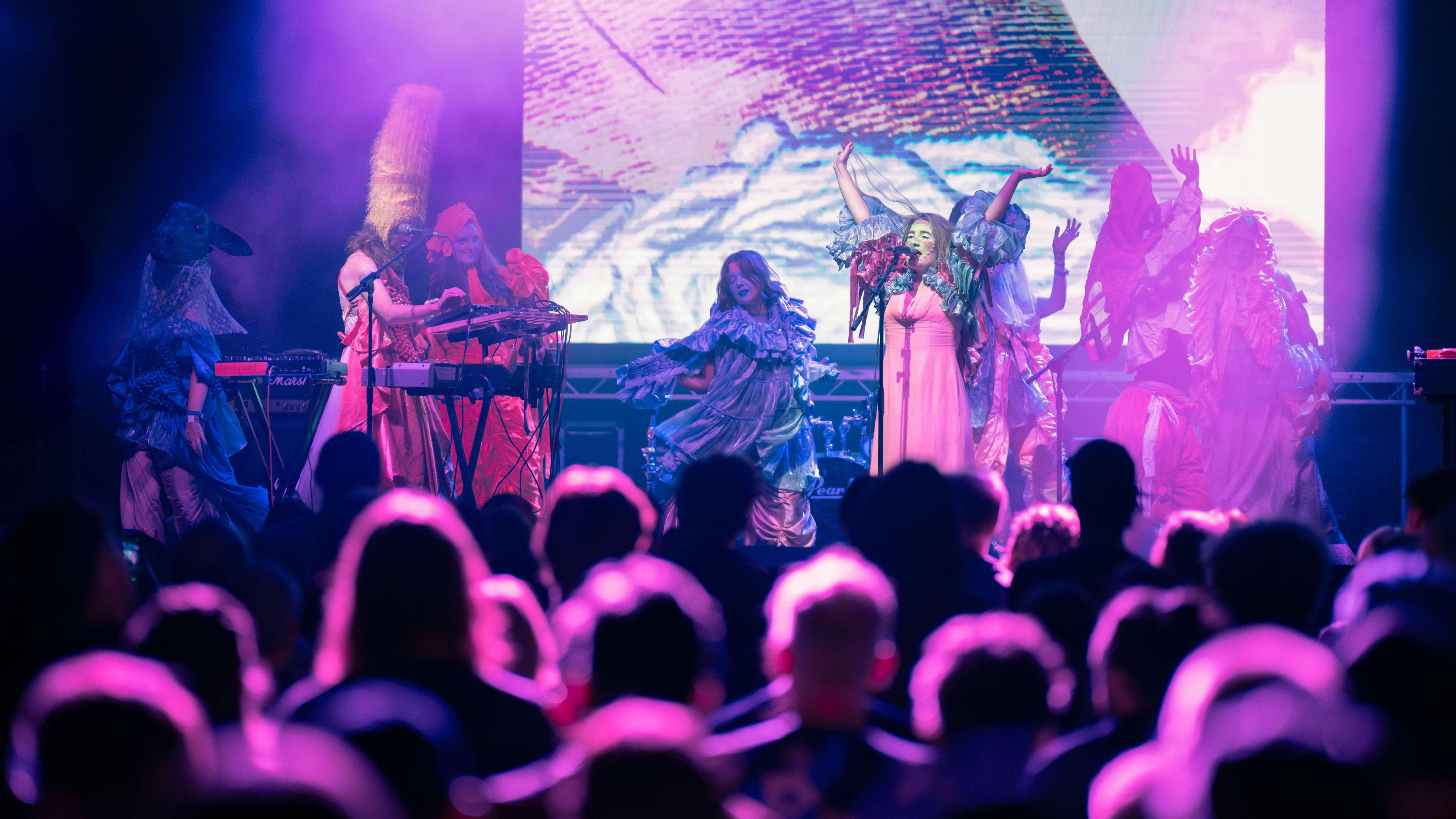 Festival audience watching Mermaid Chunky who are wearing bright costumes and hats with the stage bathed in a pink light. 
