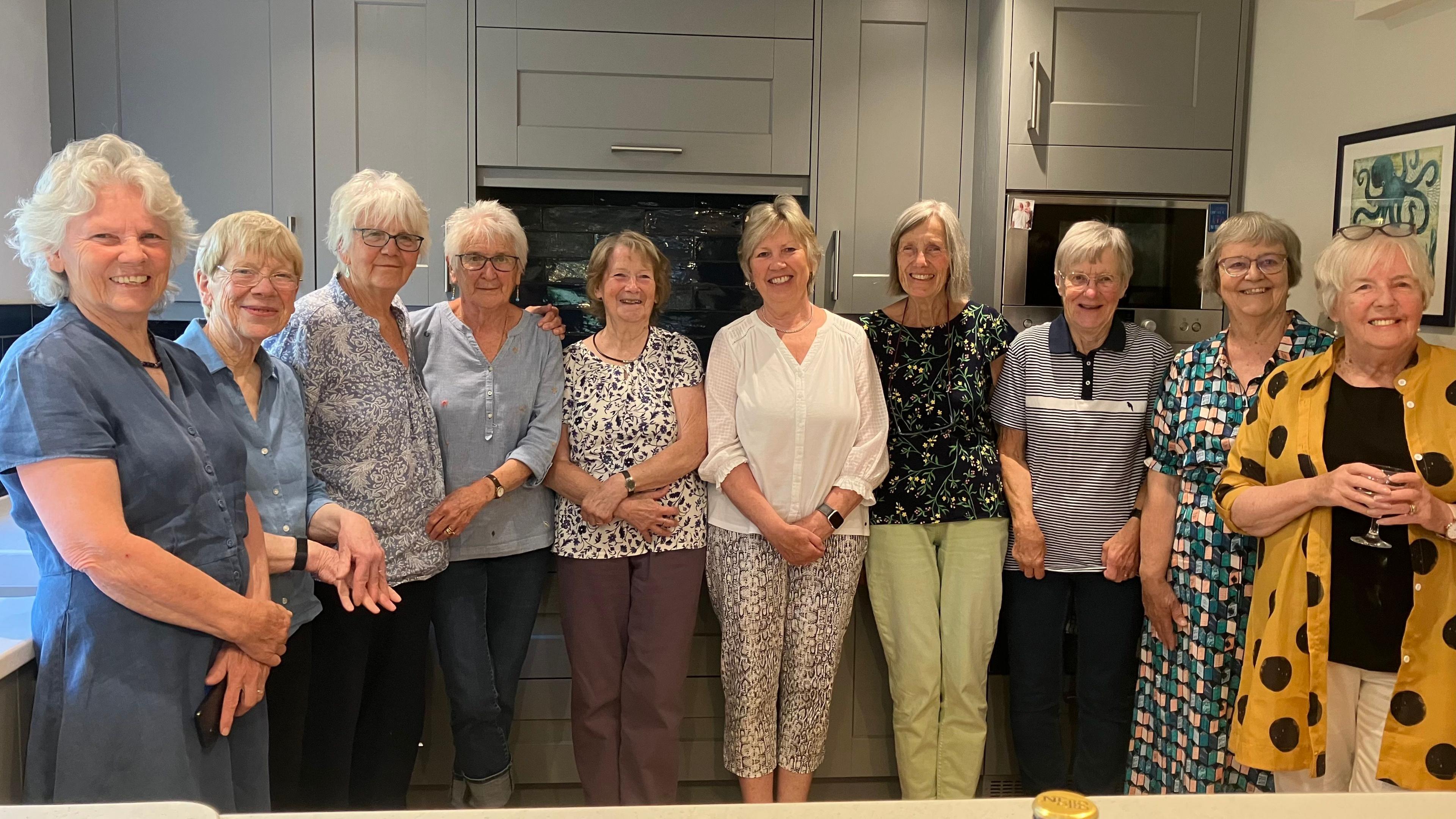 A group of 10 women in their 70s and 80s standing in a large kitchen. They are all smiling.