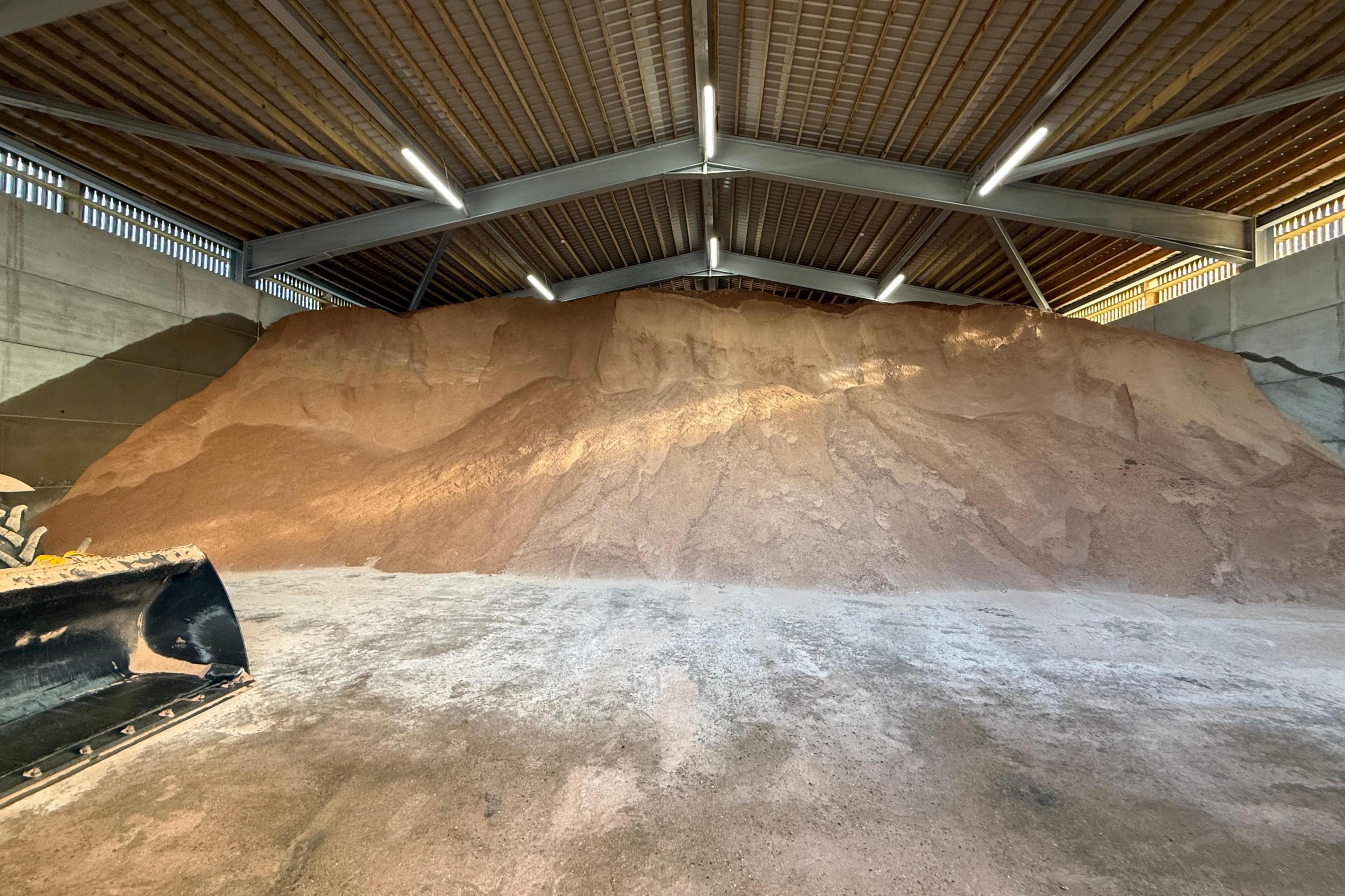 The interior of the barn, which is large and has a high roof. A huge mountain of gritting salt is inside it. Part of a digger - possibly used to scoop the salt - is to the left.