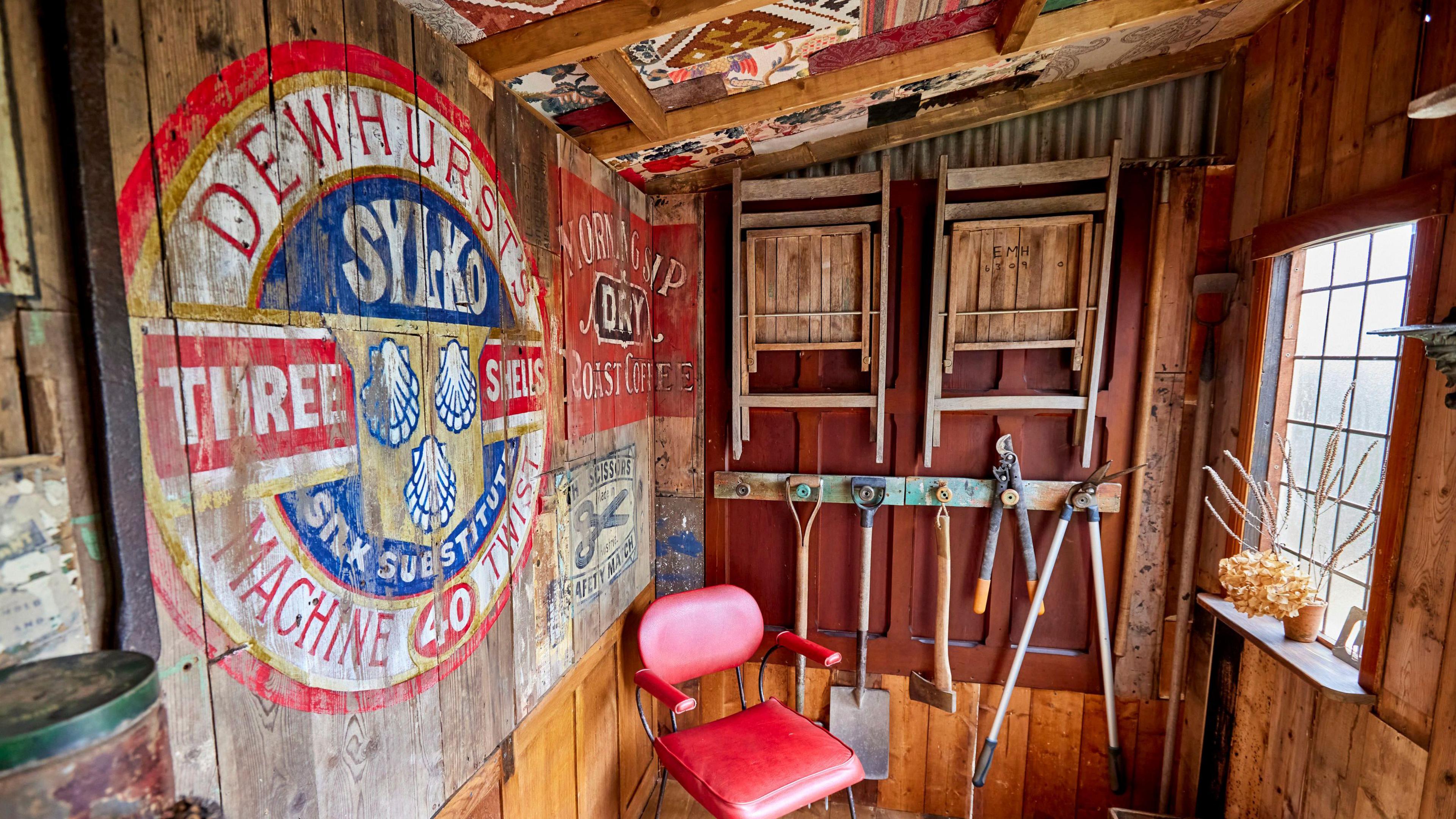 The interior of a shed with a red chair in the corner and gardening tools hung on the wall and there is a small window