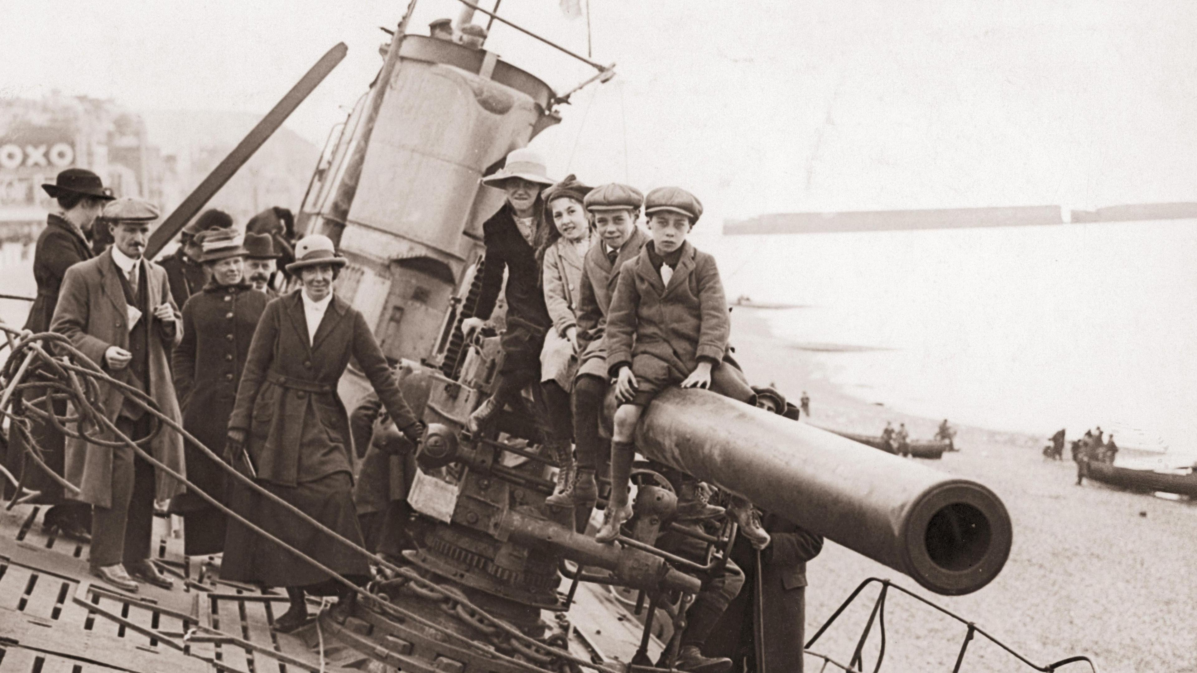 A black and white picture from 1919 shows three children and a woman sat on the barrel of the submarine's gun. To the left a crowd of men and women also smile at the camera. In the background you can see the shore of Hastings beach. 