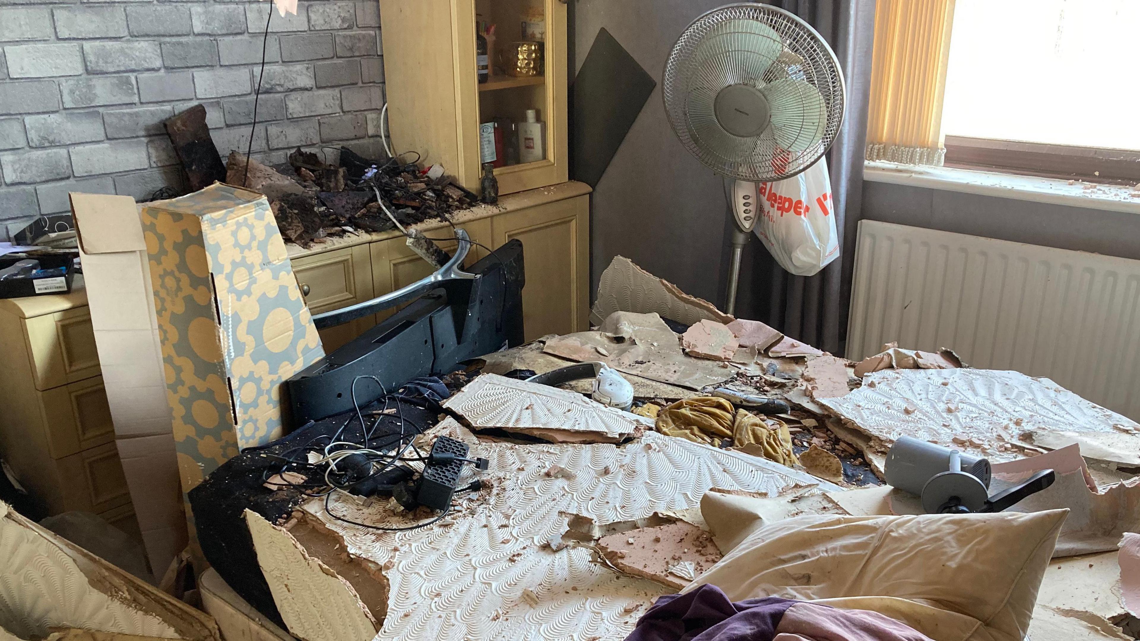 Debris on a bed in a bedroom, including wallpaper, wall plaster, rubble and electrical equipment. More detritus can be seen on a desk against the wall of the room.