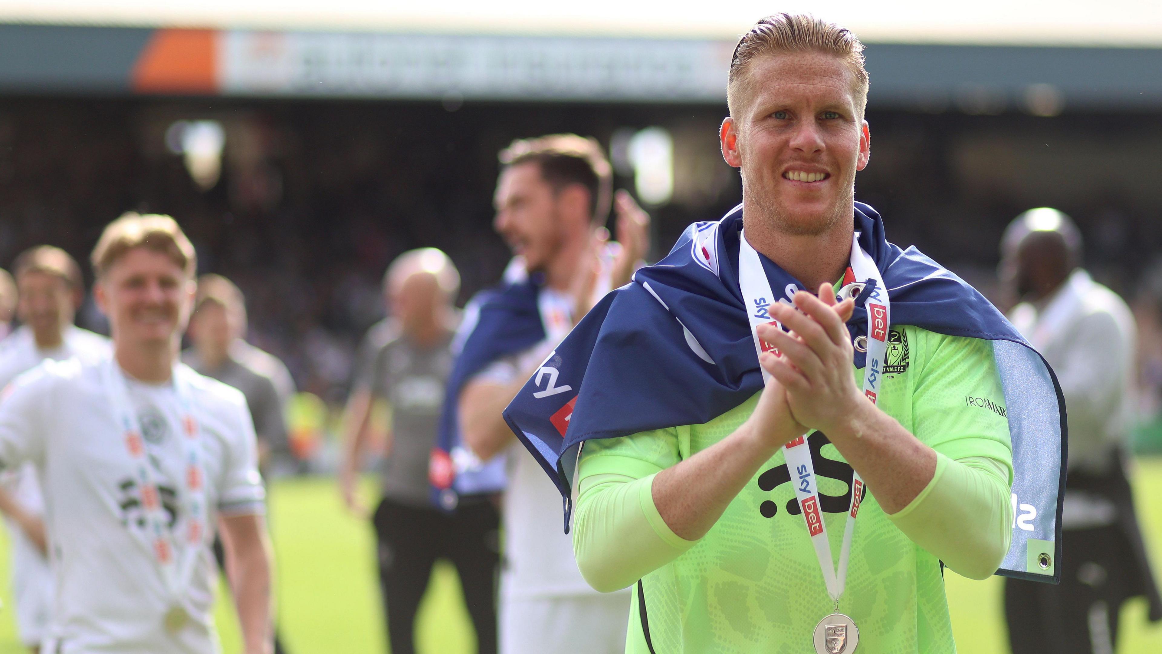 Ben Amos is wearing a League Two promotion medal around his neck and a blue flag over his shoulders as he applauds the Port Vale fans after the club win promotion