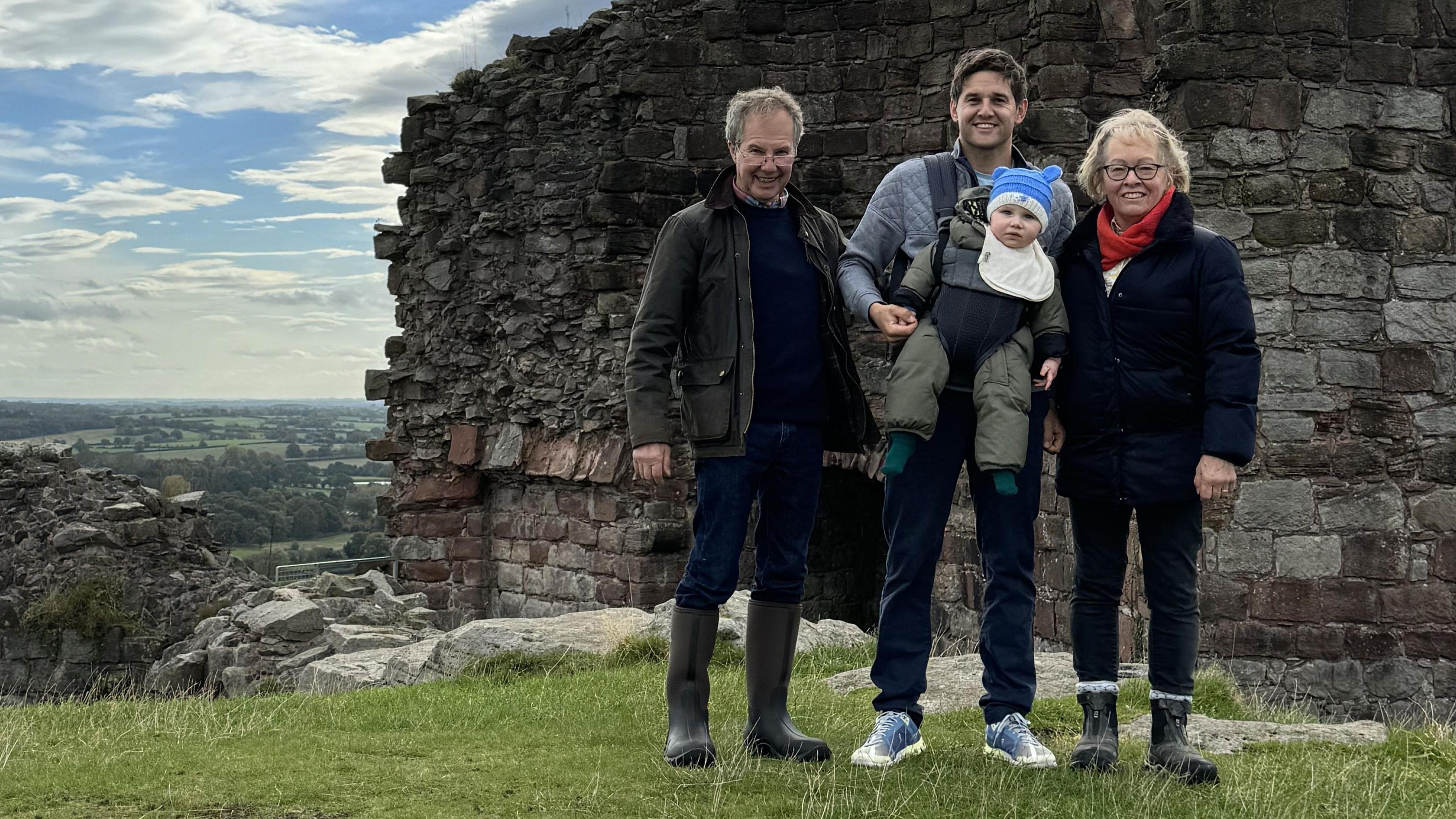 Grŵp teuluol gydag Alex Loven yn y canol, yn cario'i blentyn ifanc. Mae'r grwp yn sefyll yn yr awyr agored, gyda chastell a golygfeydd pert y tu ol iddynt.