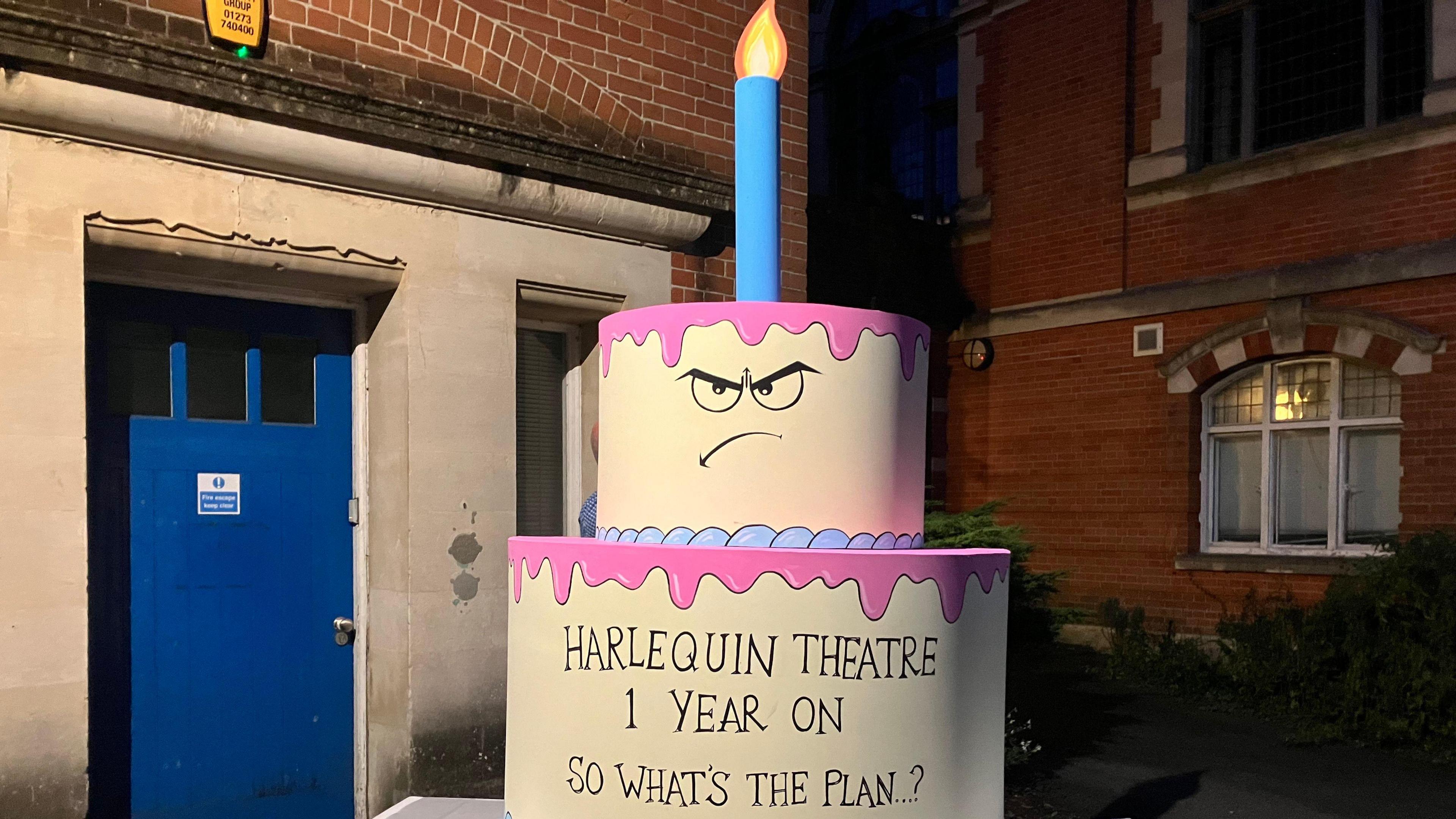 A model birthday cake with two tiers and a single cake on top. the top tier had an angry face drawn on it while the bottom tier reads Harlequin Theatre one year one So what's the plan?