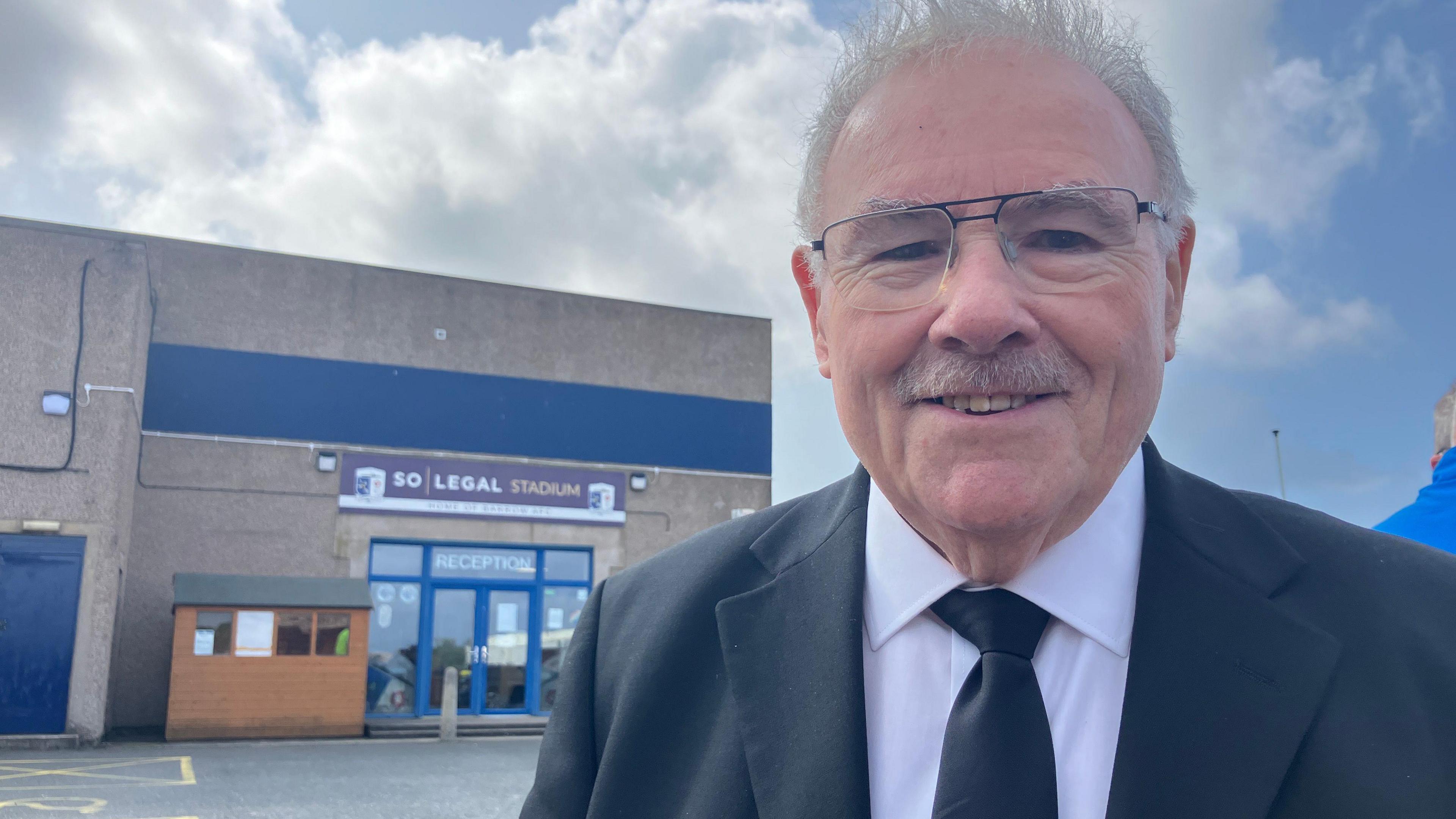 Mr Herbert has short white hair, a moustache and glasses. He is wearing a black suit and tie and looking into the camera.