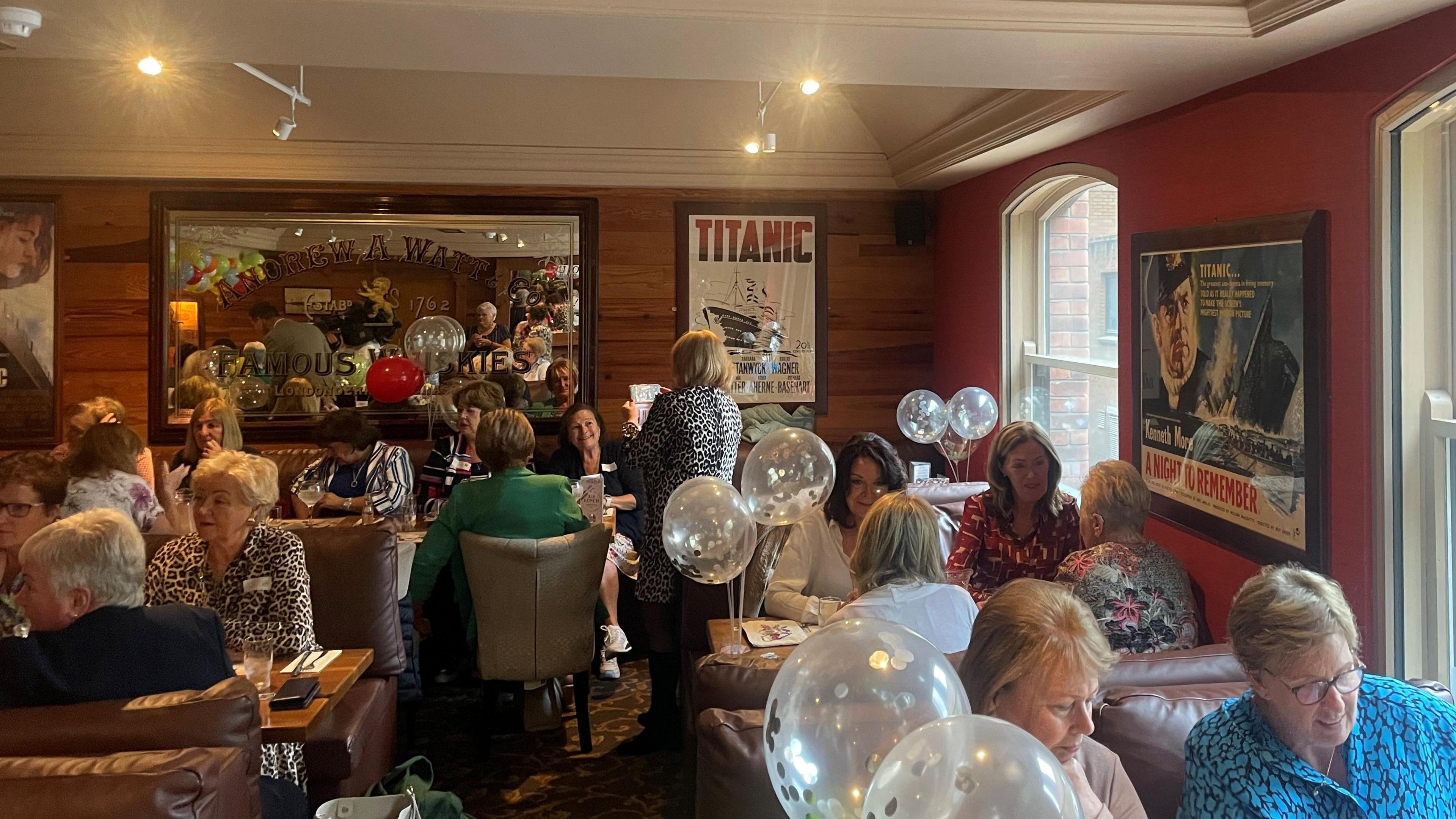 An array of nurses at the event, there are multiple balloons and titanic themed posters hanging on a wooden wall, all the ladies are engaging with one another in conversation