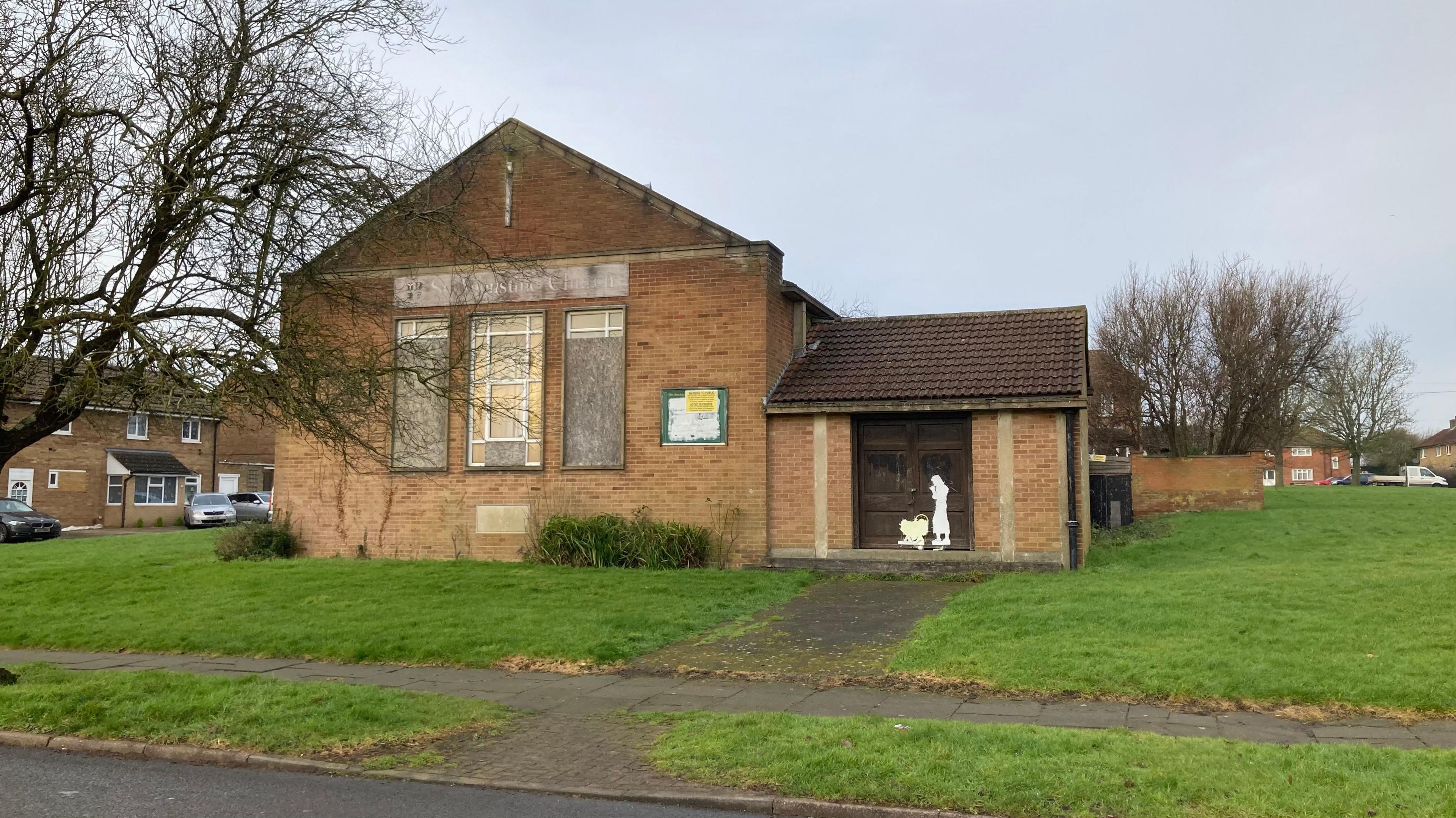 Brick church building with boarded-up tall windows and a small extension to the right. A Christmas silhouette is visible on the extension. It is located in a grass square surrounded by houses. 