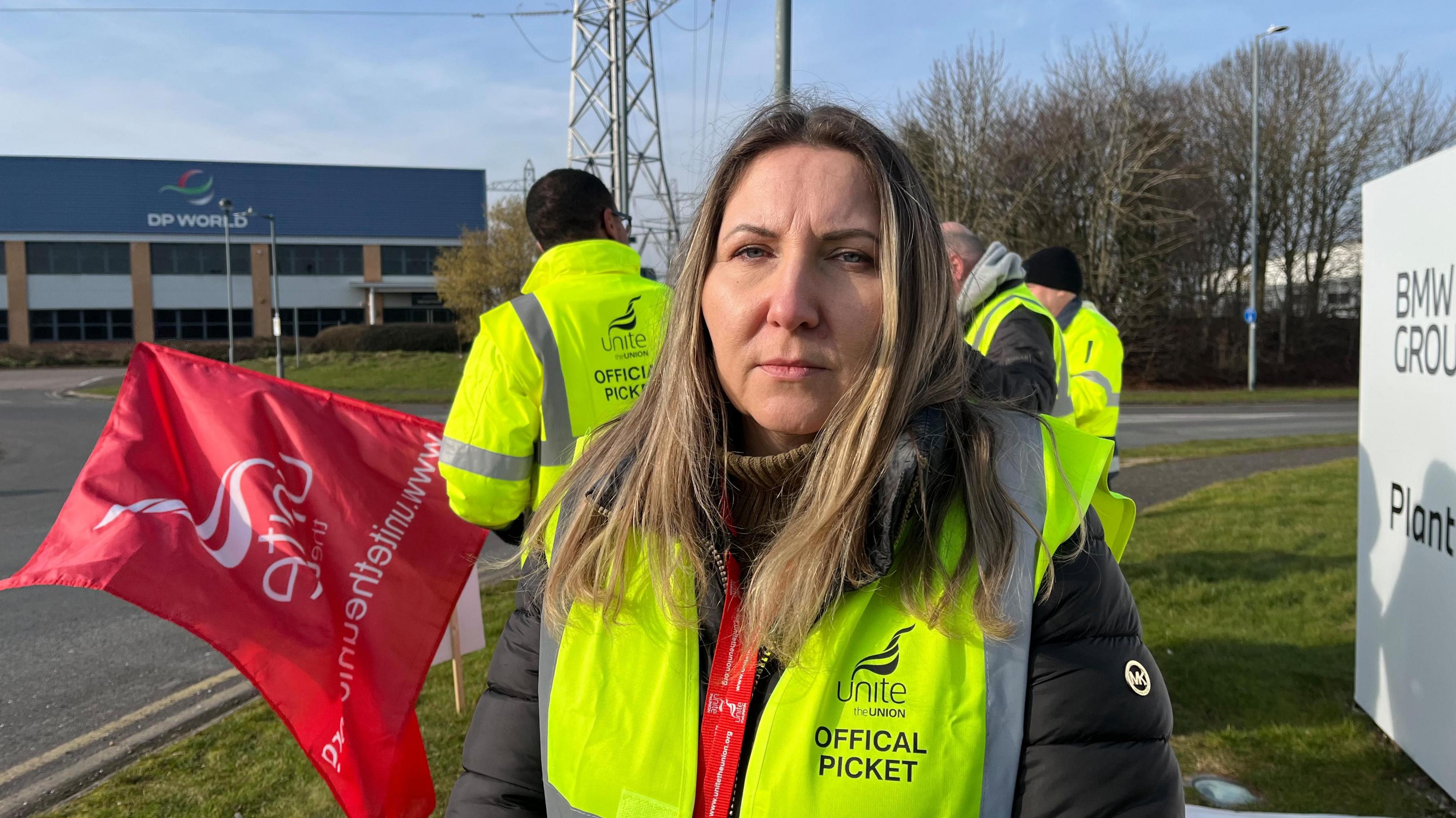 A woman with blonde hair and a high-visibility yellow jacket looks at the camera as she stands on the picket line outside the BMW Hams Hall plant in Coleshill.