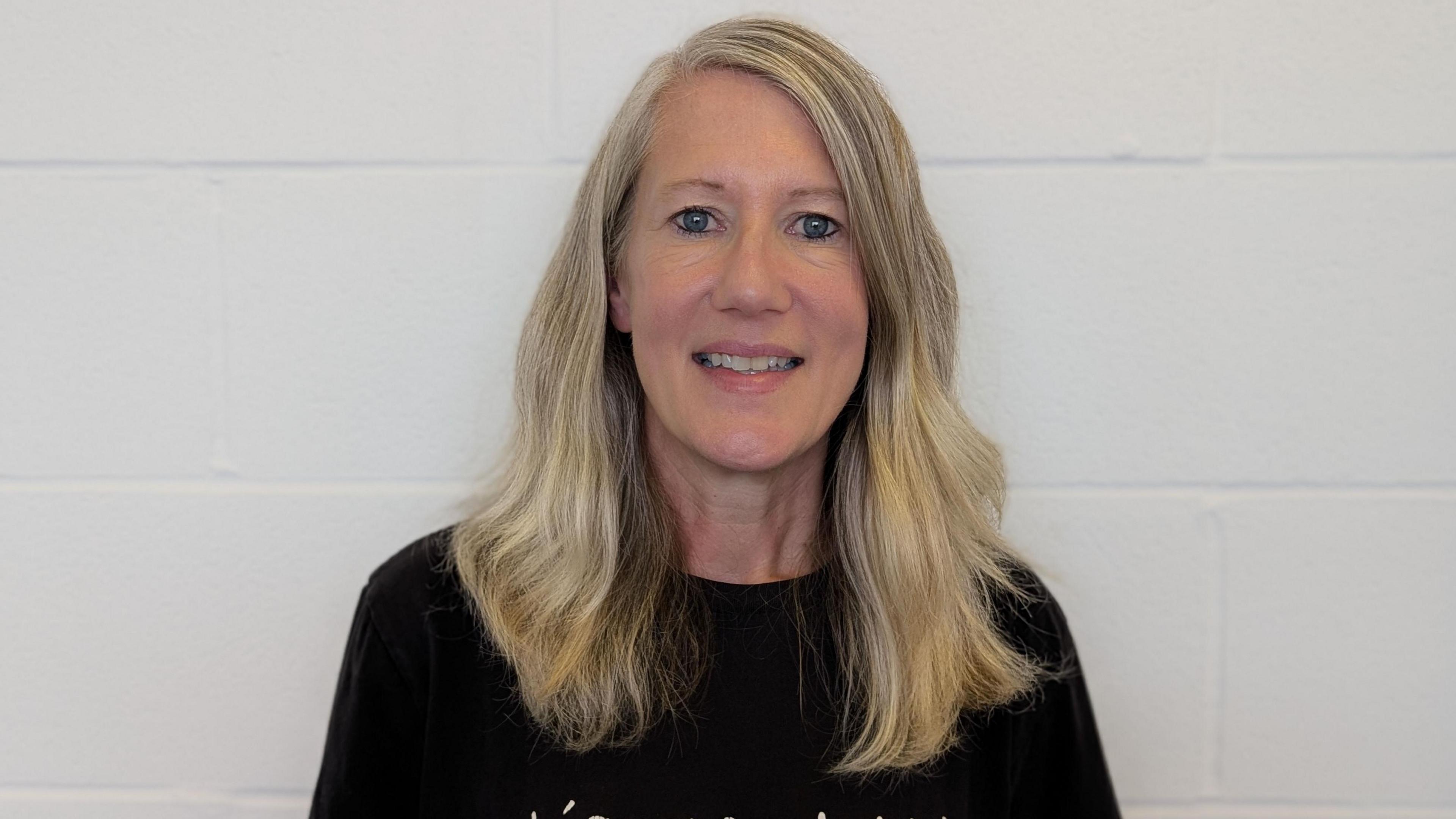 Rhiannon has long blonde hair and wears a black t-shirt. She's smiling at the camera against a white brick wall.