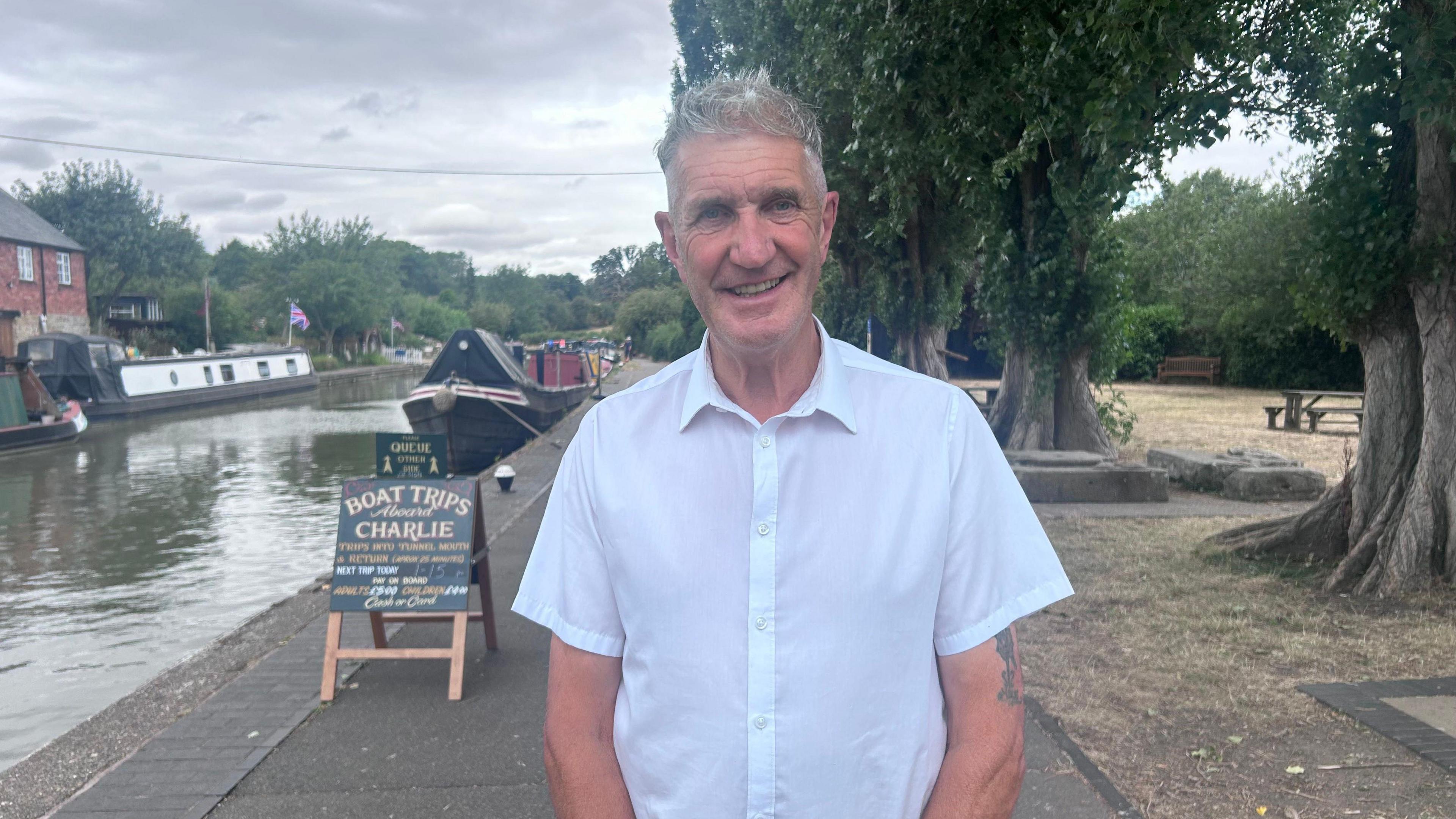 A man with short grey hair and  wearing a white short-sleeved collared shirt, is standing on a canal-side path next to an A-board sign advertising 