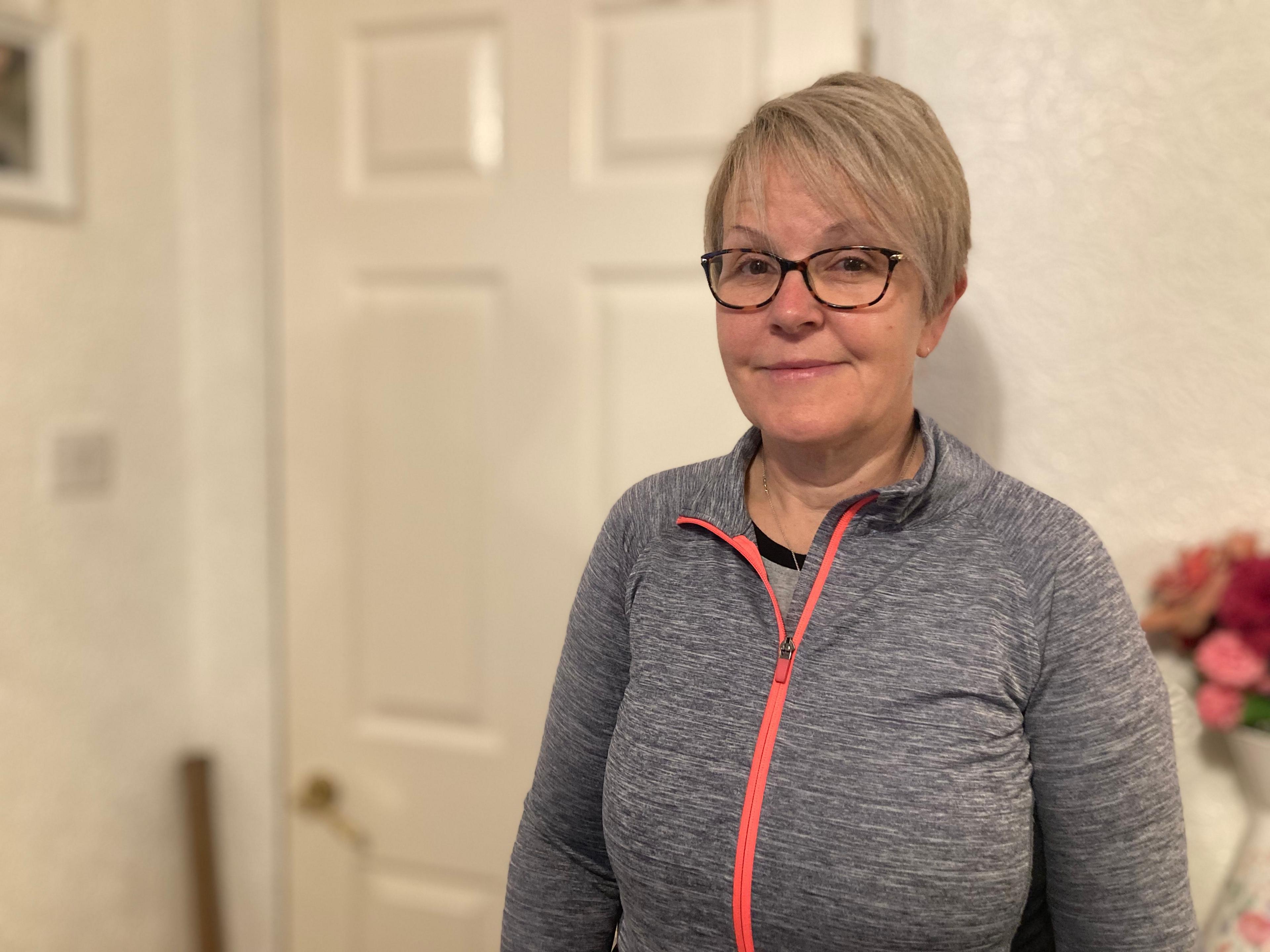 A woman with short blonde hair wearing glasses and a grey jacket stands inside a house, behind her is a white door, white walls, and a partially visible vase of red and pink flowers