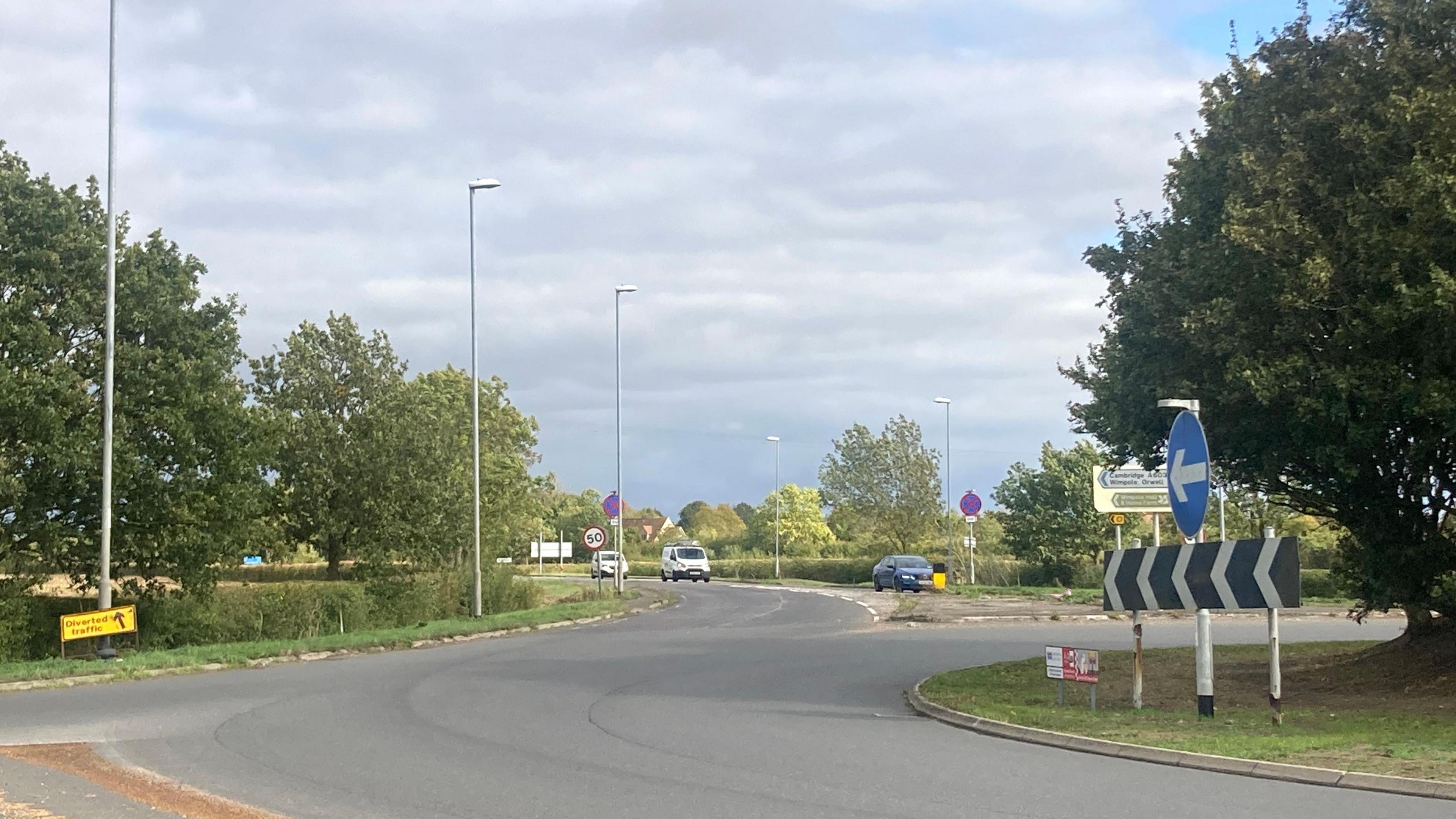 Part of a roundabout. Road signs detail the direction of travel and nearby places. Cars can be seen in the distance approaching the roundabout.