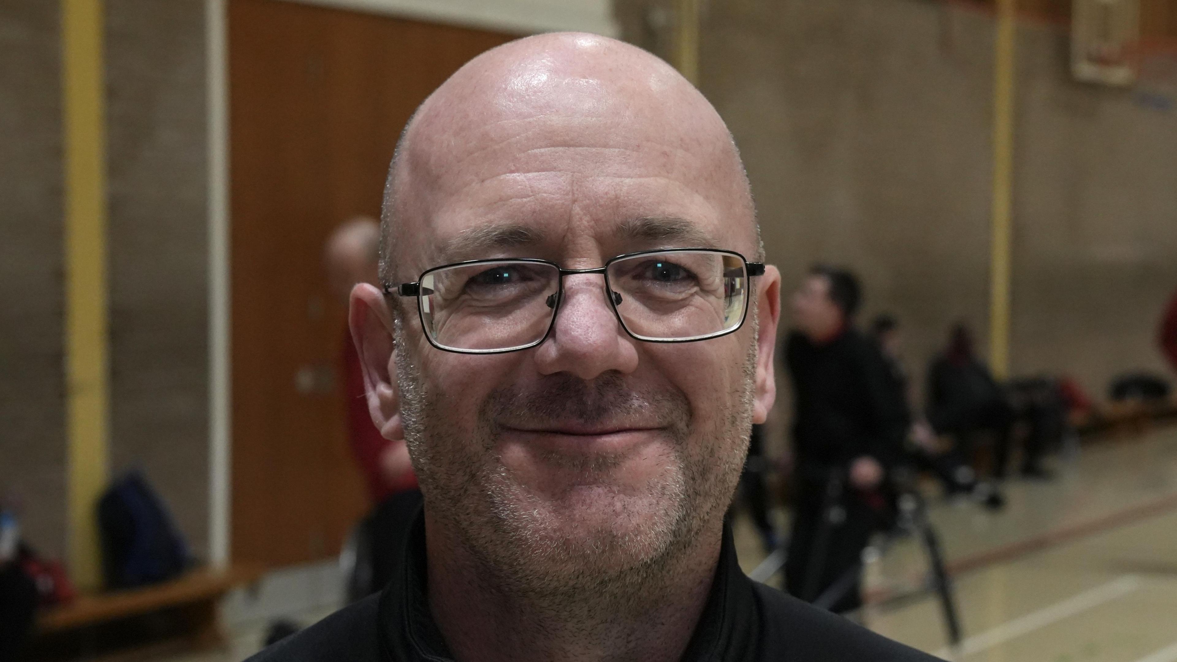 Ian Ling smiles at the camera while standing in a sports hall. He is bald and wearing glasses. He is wearing a black jumper. Some club members can be seen pictured behind him talking with each other.