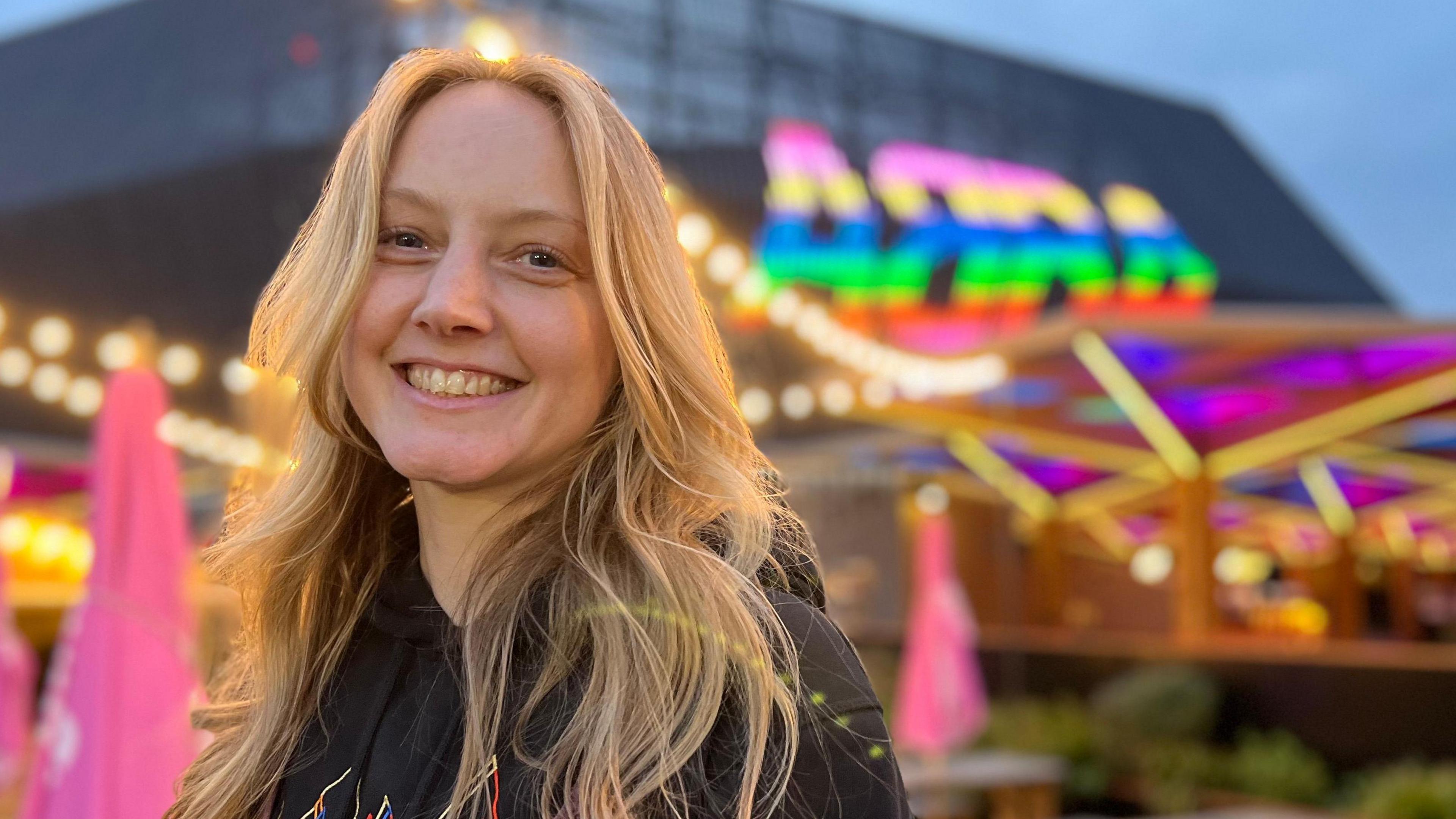 Janette, who has long blonde hair and wears a black hoodie, smiles outside the Abba Arena, which features the word 'Abba' in giant multi-coloured lights on the outside.