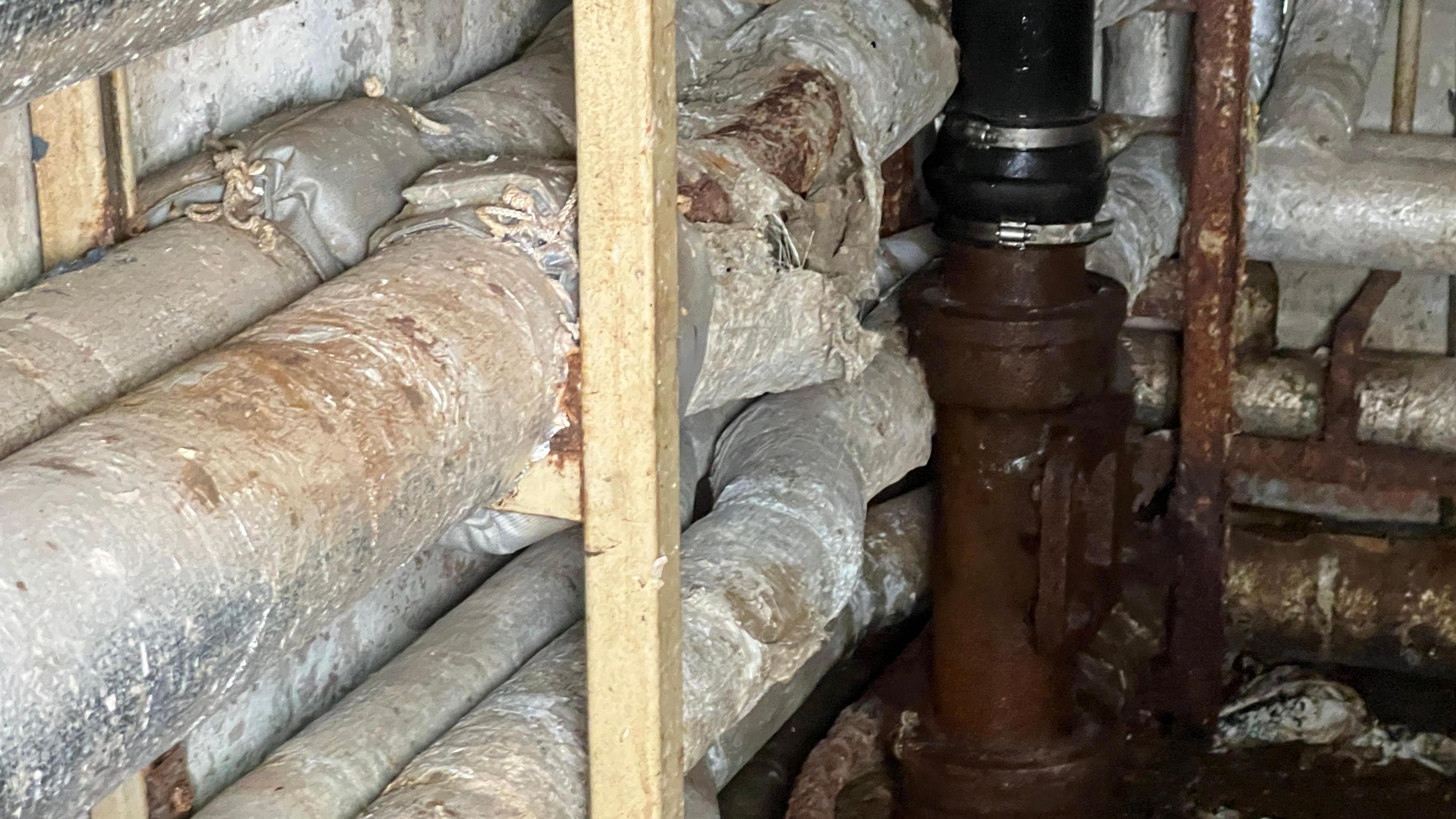 A series of pipes, some cladded with silver material; some exposed and looking rusty. The is a wooden upright next to them.