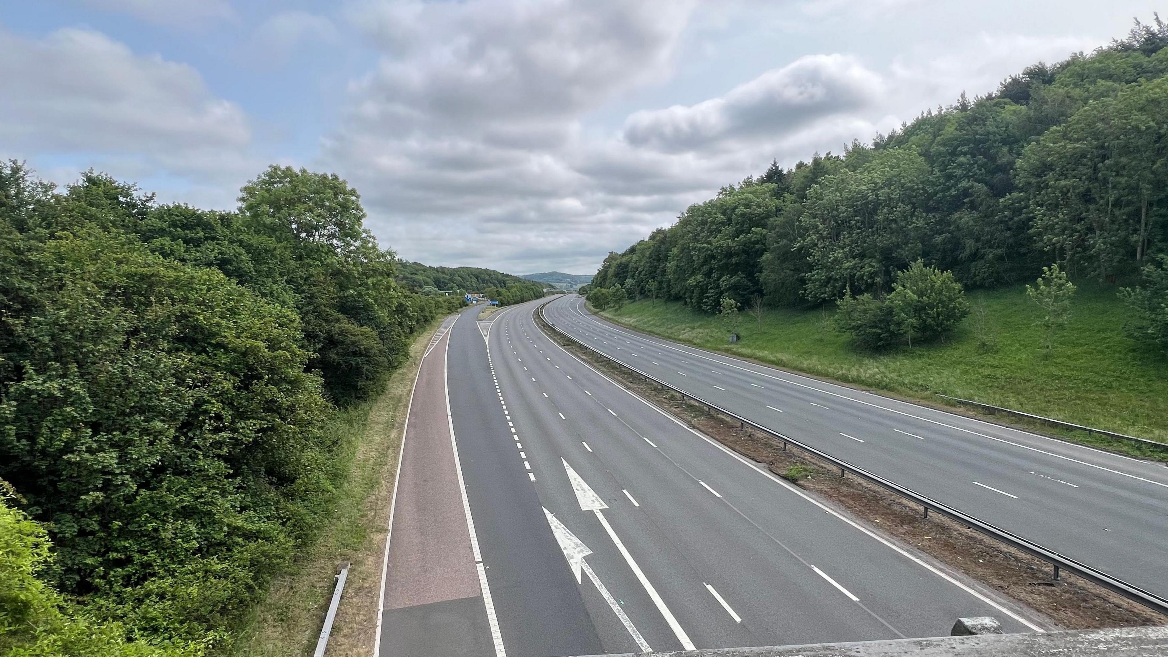 The M5 motorway completely clear of cars in both directions. There are grassy banks and trees on either side. The sky is overcast with areas of blue peeking through.