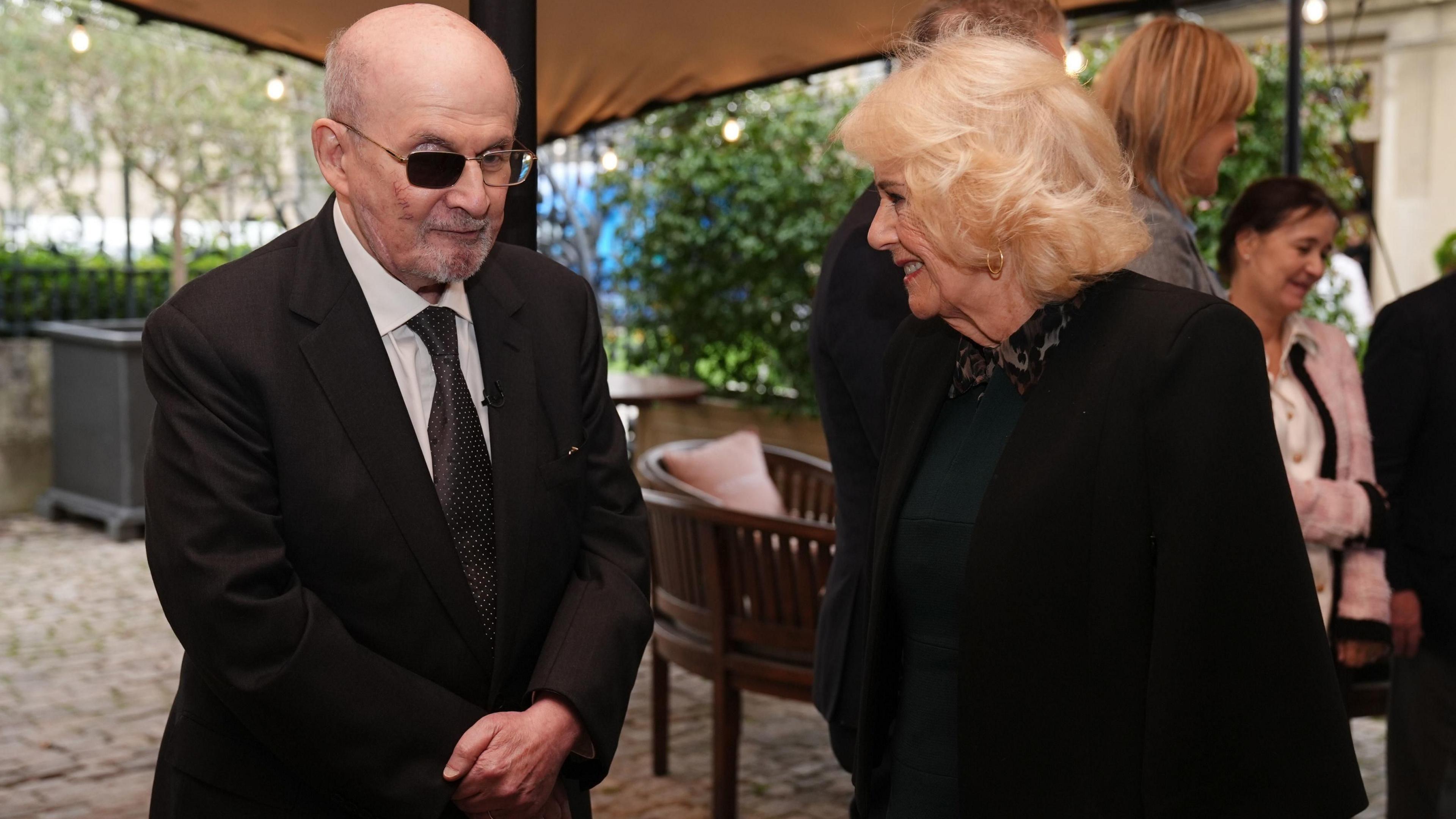 Queen Camilla speaks to Sir Salman Rushdie, who is wearing a dark suit and glasses with one blacked out lense, concealing an injury he sustained when he was attacked on stage two years ago.