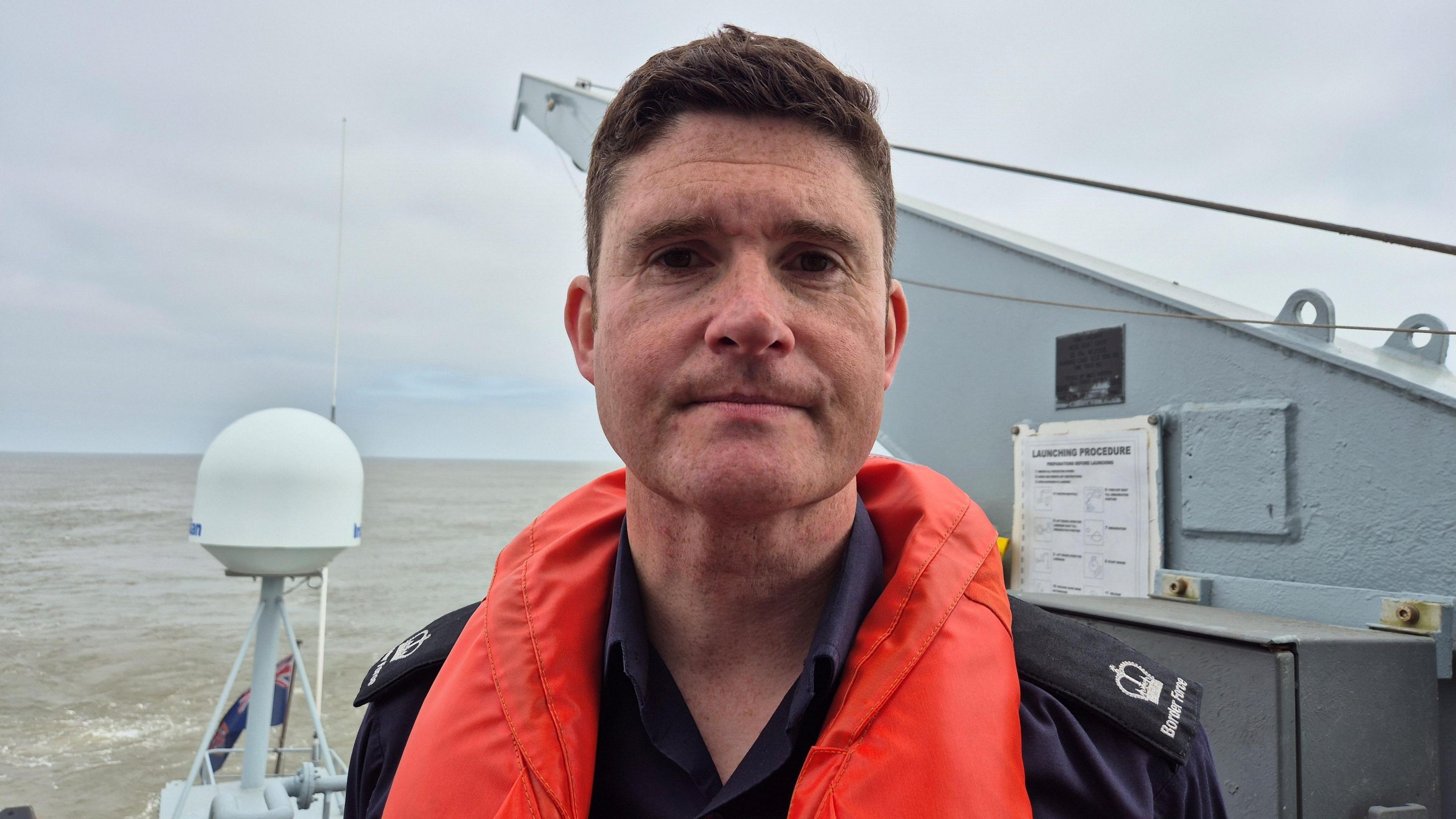 Andy Vidamour is standing on a grey boat. He has short brown hair and is wearing a navy blue shirt with crown logos on lapels. He is wearing an orange life jacket.