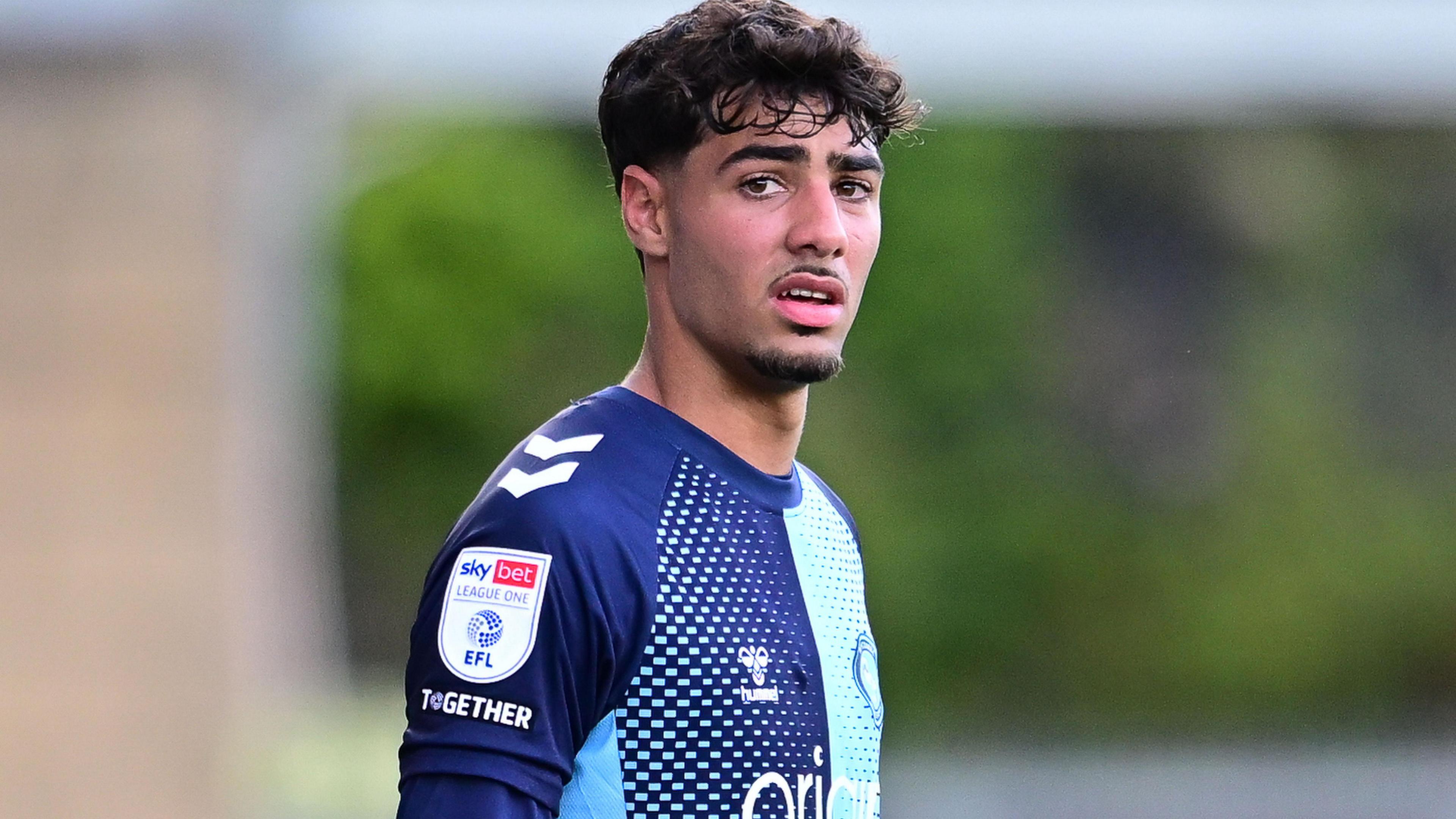 George Abbott hace una mueca mientras mira a la cámara. Viste la equipación azul claro y oscuro del Wycombe.