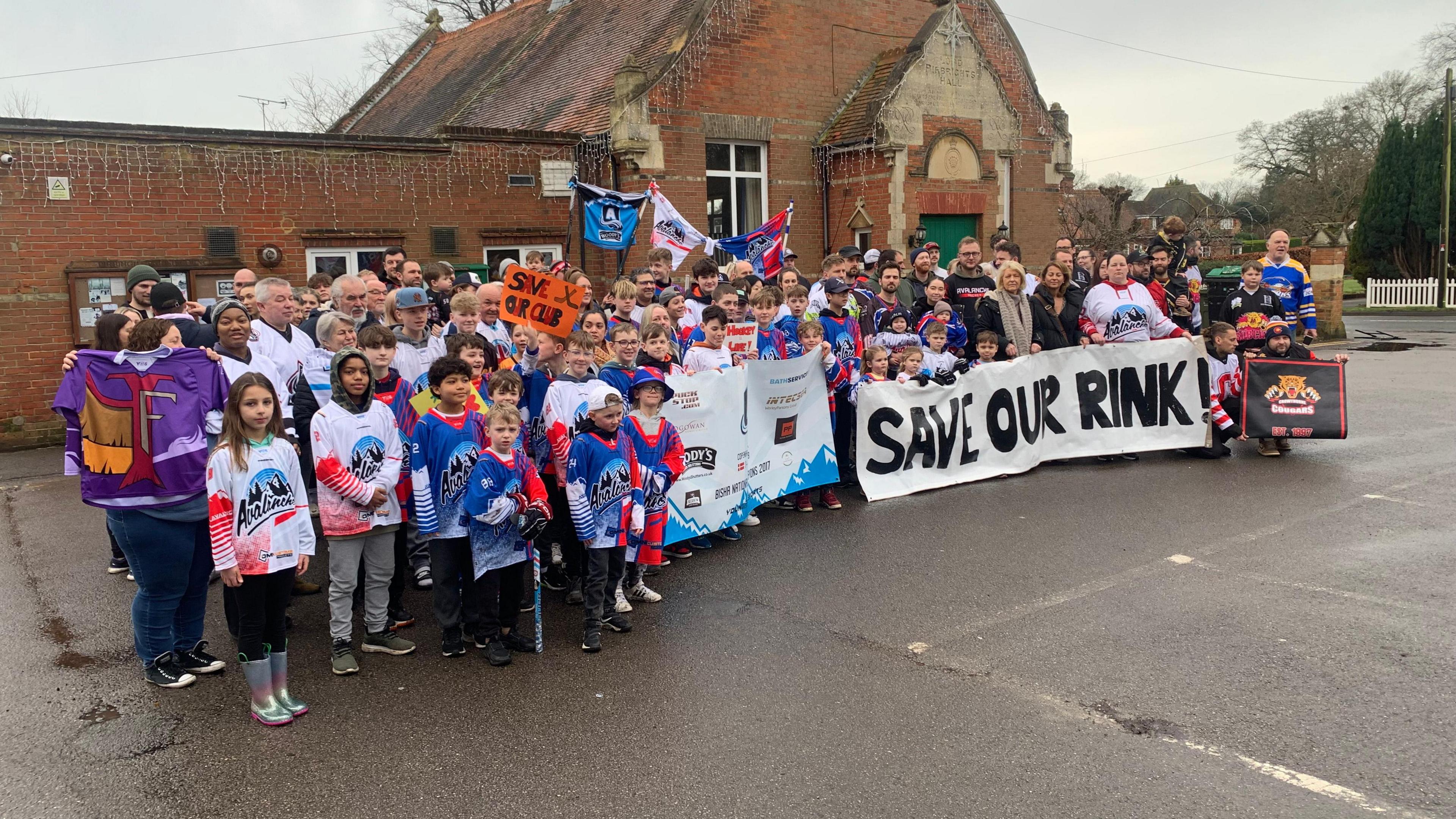 A group of hockey players of all ages wearing multi-coloured hockey jerseys. They also have a sign reading "Save Our Rink!".