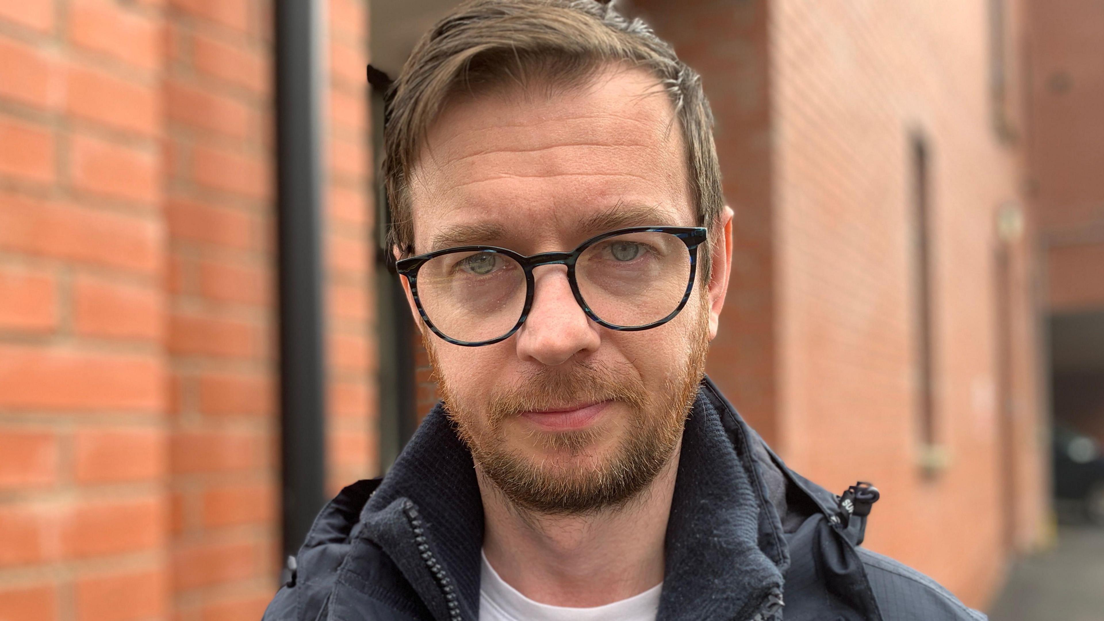 A man with short dark hair and dark facial hair is speaking as the picture is taken. He is wearing black round glasses and is standing in front of a red brick building. He is wearing a navy coat with a pink zipper and white t-shirt. 