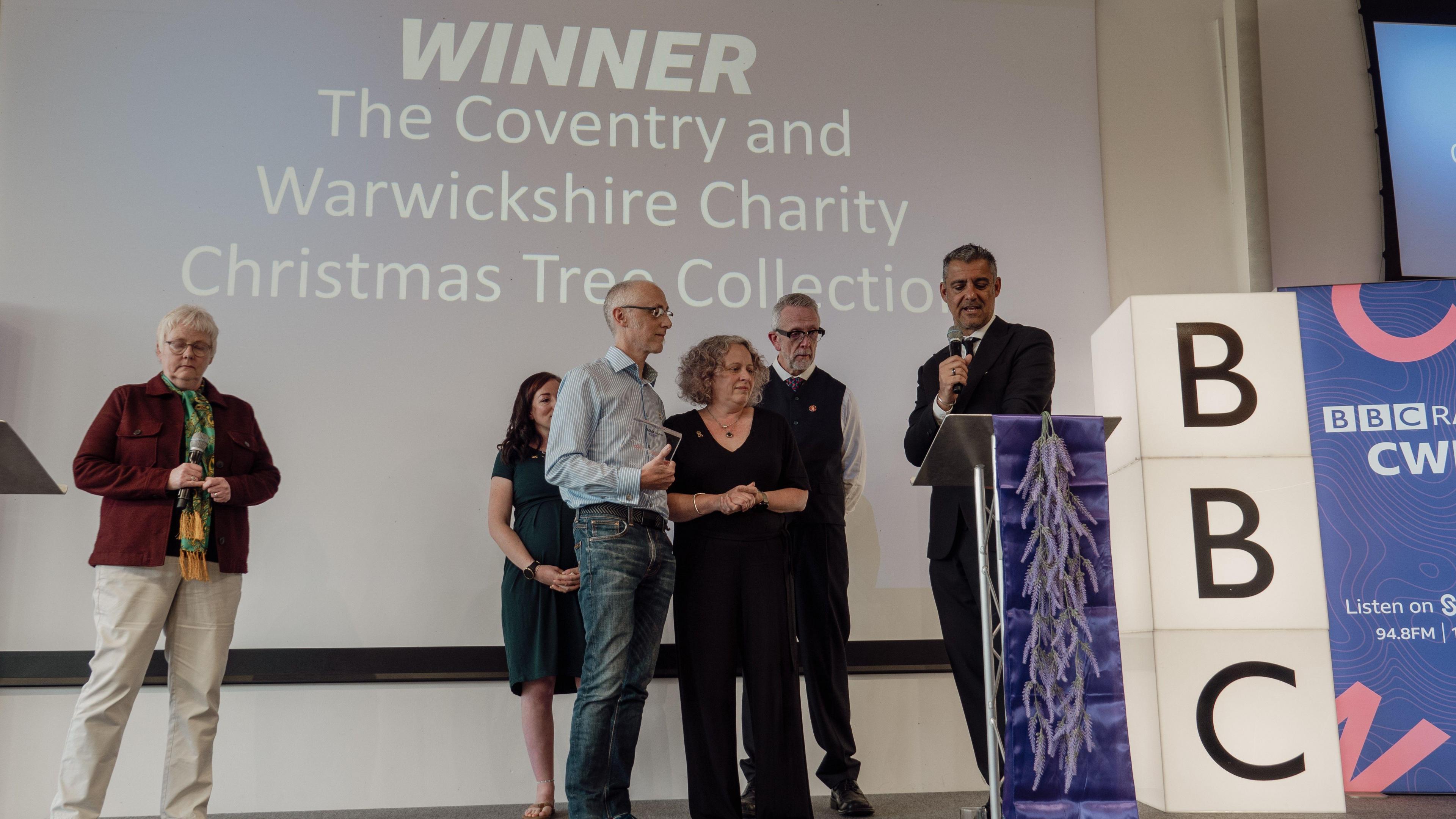 Three people collect an award on stage. There is a man standing next to them with a microphone, a woman standing behind them, and another woman standing to the side. There is a purple plinth, and a large light-up BBC sign.