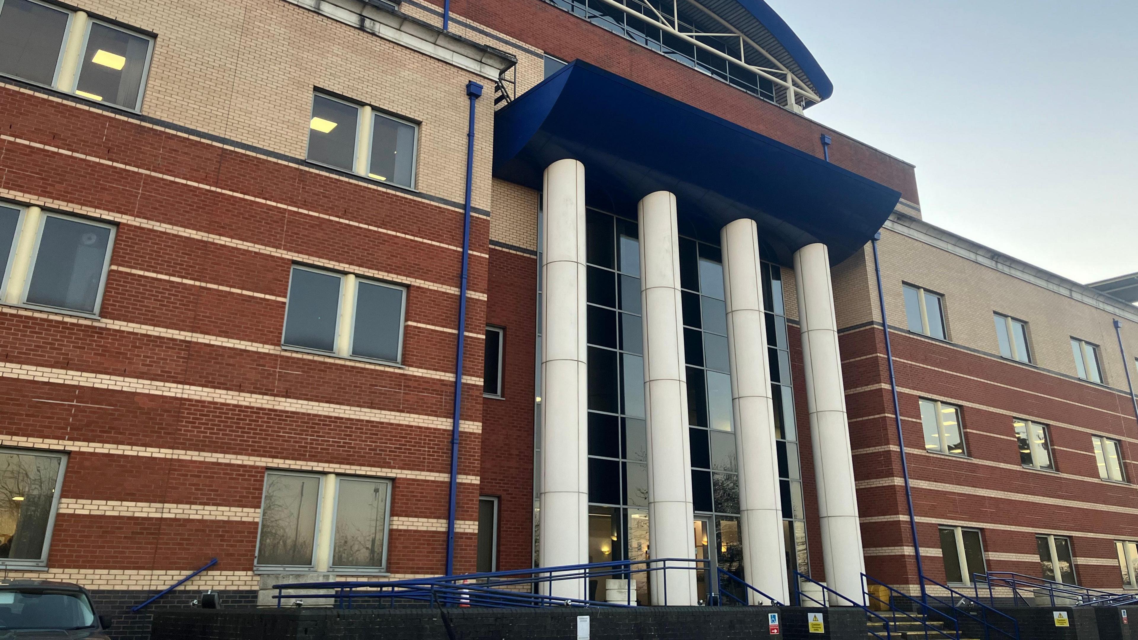 A three tiered red brick building with a glass windowed entrance with four white columns at the front.