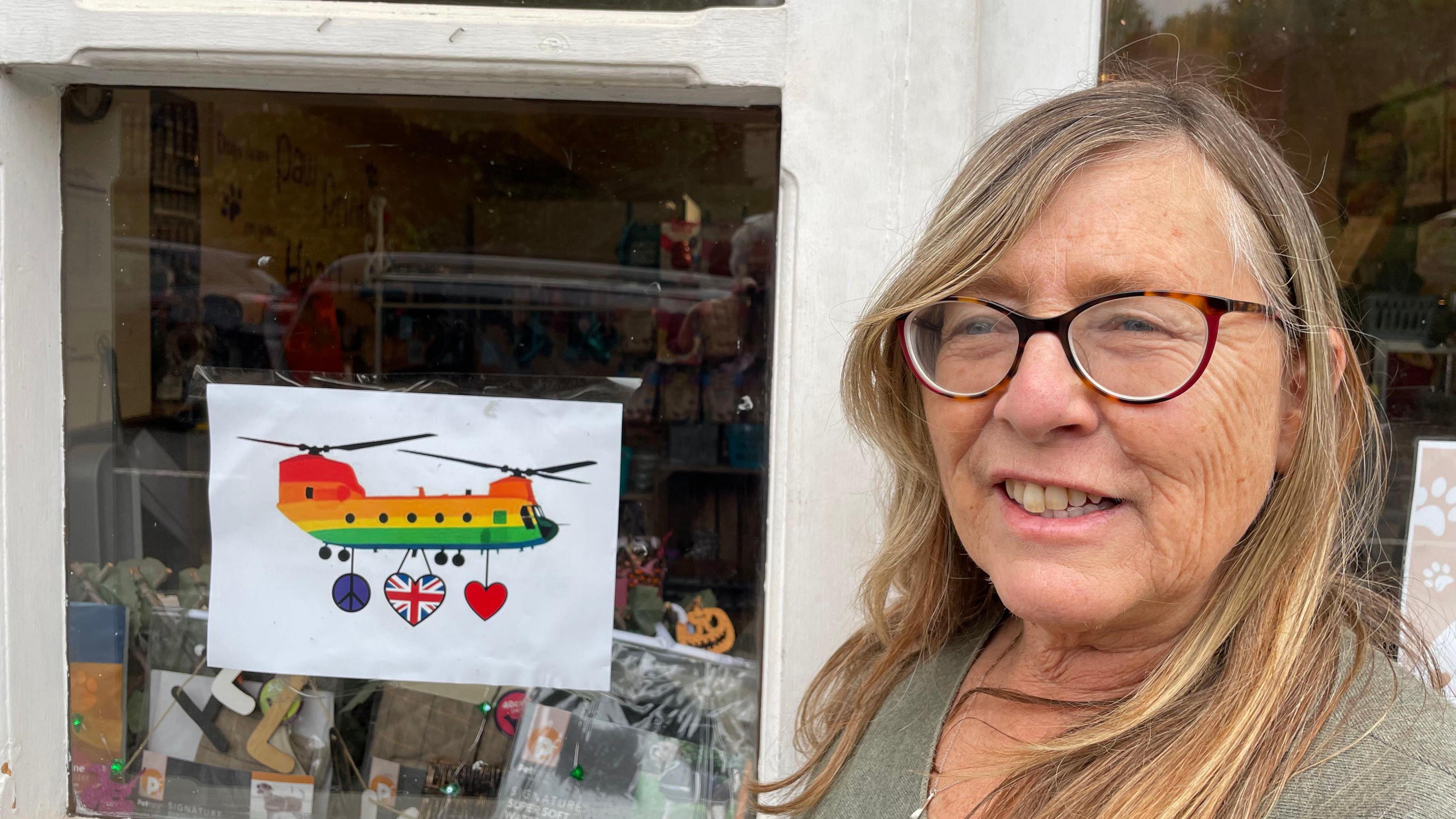 Woman with long hair and glasses in front of a shop window featuring a picture of a brightly-coloured helicopter