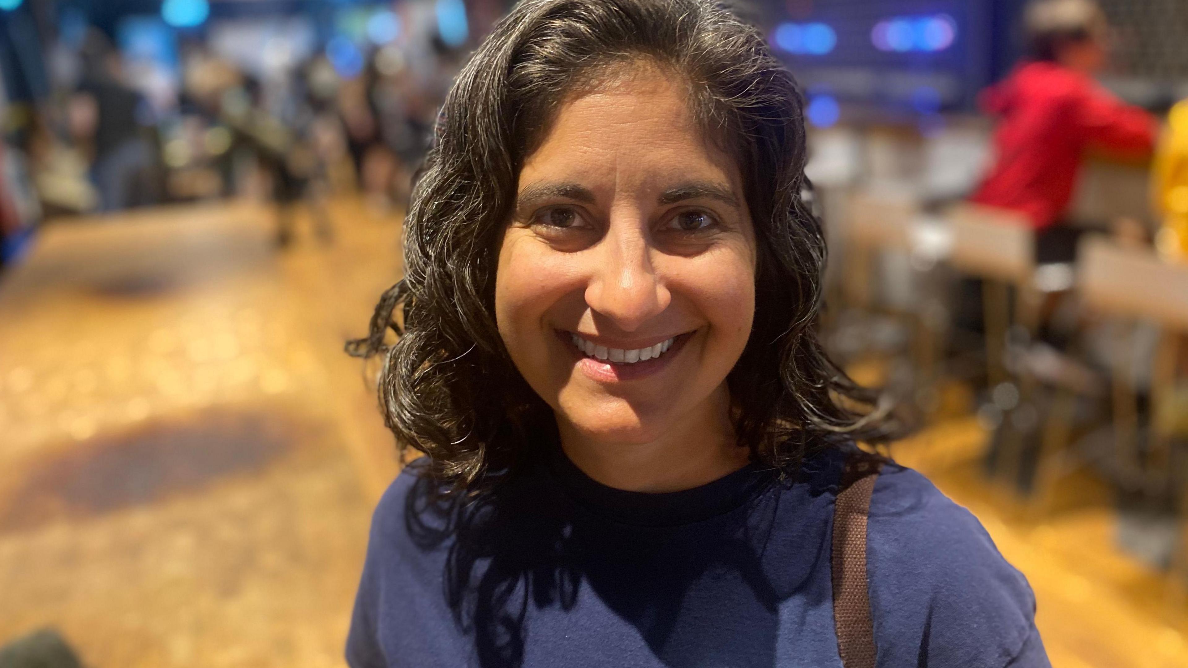Nicky Shepard who is smiling at the camera an wearing a blue sweatshirt. She has curling shoulder-length black hair. Behind her is a blurred out hall and just glimpsed is a row of desks and chairs, two with the back view of young people standing up and reaching across the desk.