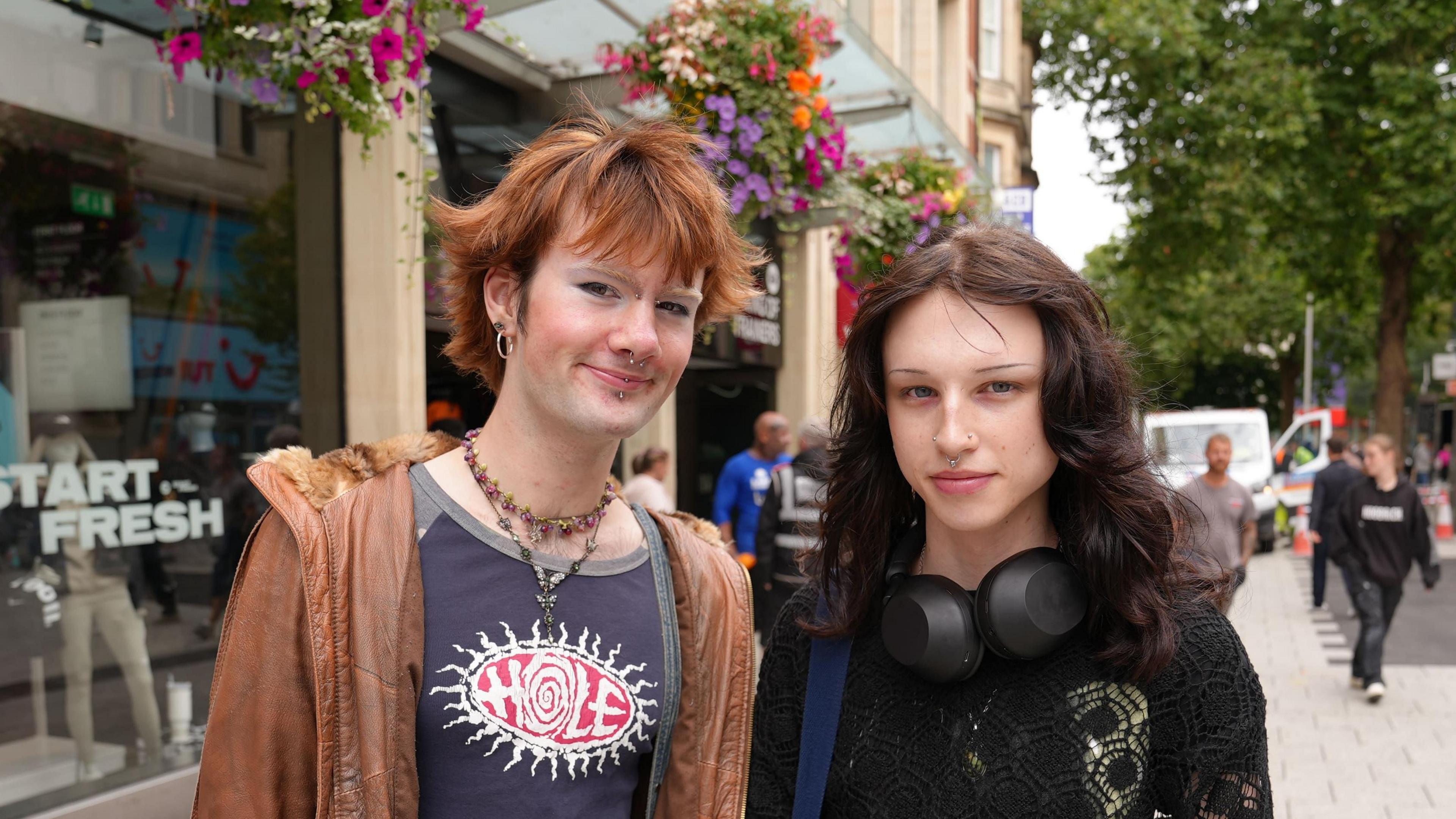 Leo Lee-Thomas on the left with ginger slightly spiked hair. They are wearing necklaces and a navy top that says 'hole' with a sun around it. They are wearing a brown leather coat. Next to them is Aiden Forrester who has long brown curly hair, with their headphones around their neck and a crochet jumper