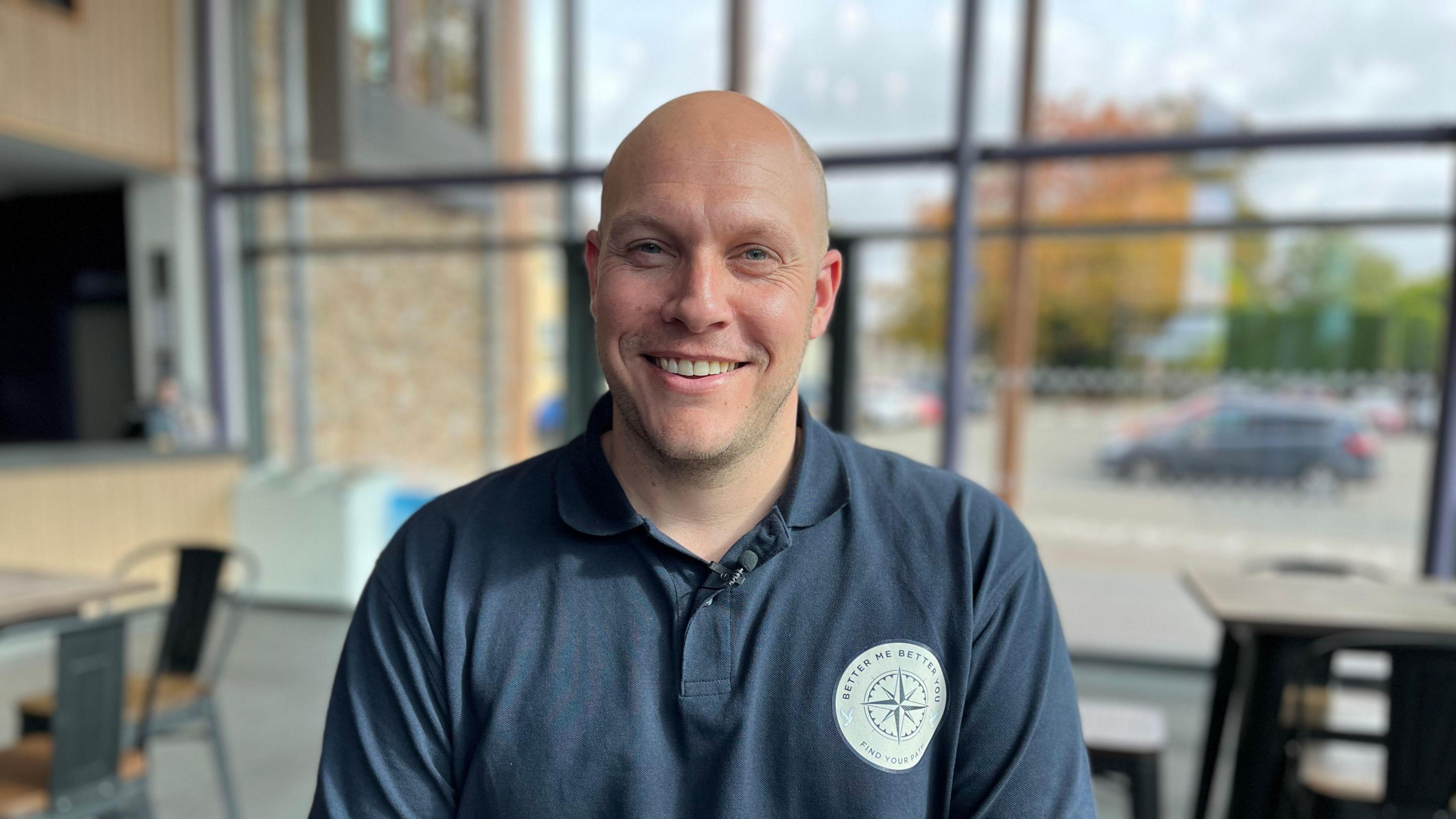 Sam Coton wears a navy polo shirt with the 'Better Me, Better You' logo on it. He is sitting in the school theatre bar with floor to ceiling glass windows, you can see tables and chairs behind him. He is smiling at camera.