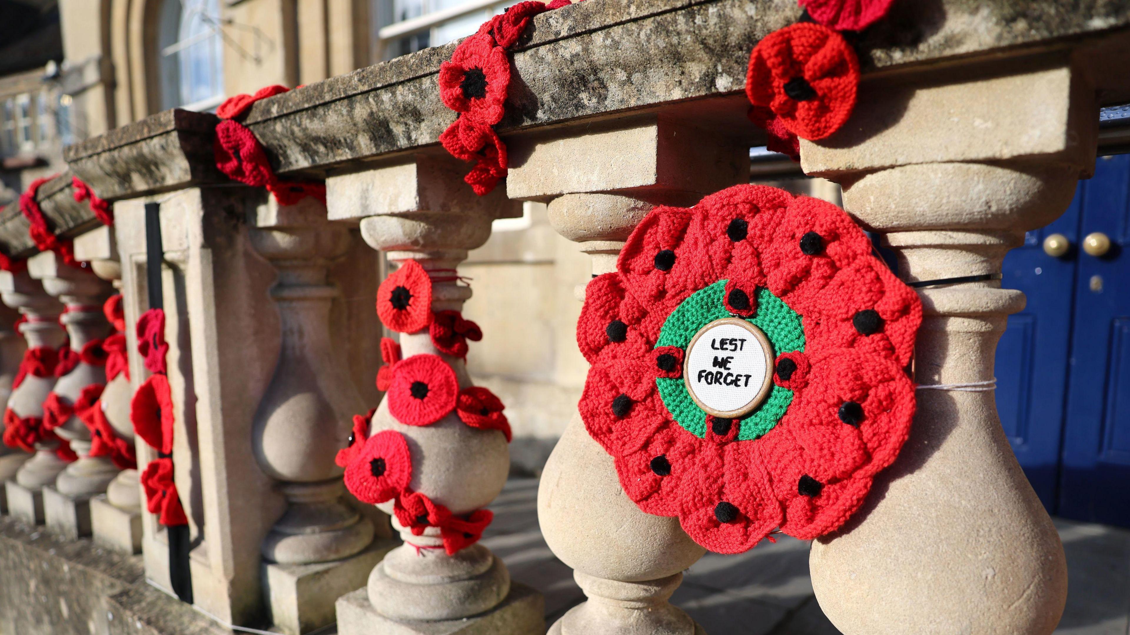 A red knitted wreath with poppies around the outside and 'lest we forget' on a white circle inside