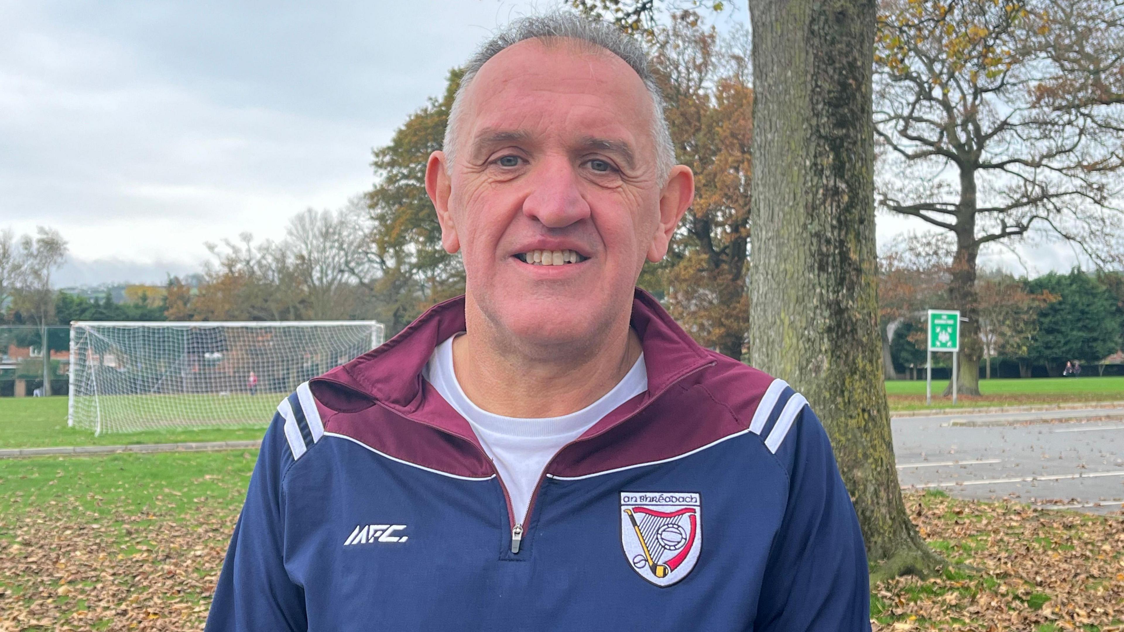 John Deery has short grey hair and is wearing a tracksuit top which is mostly navy blue but is purple around the neck and shoulders, underneath it is a white T-shirt. He is standing outside, behind him is an oak tree and further back a set of football nets.