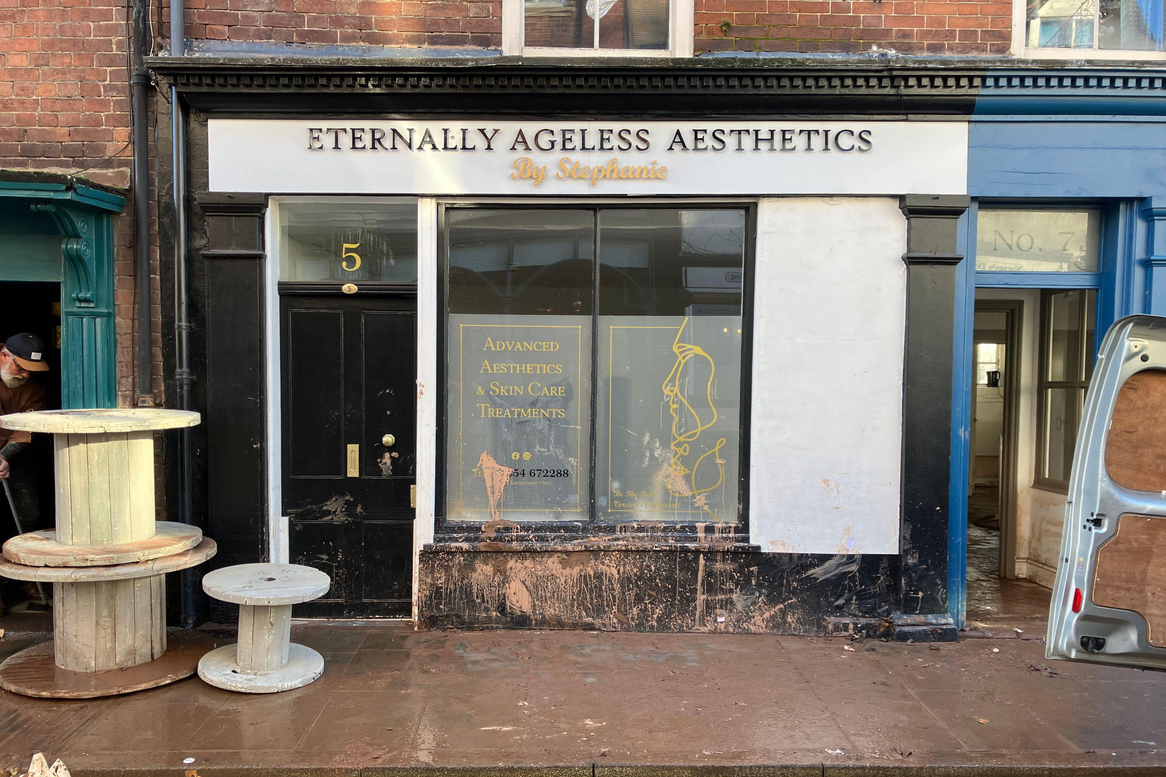 Eternally Ageless Aesthetics with black and white frontage covered in mud and flood water. Several large wooden spindles, used to wrap rope or wire around, sit outside the building. A number 5 is above the black door and there is yellow writing on the windows.